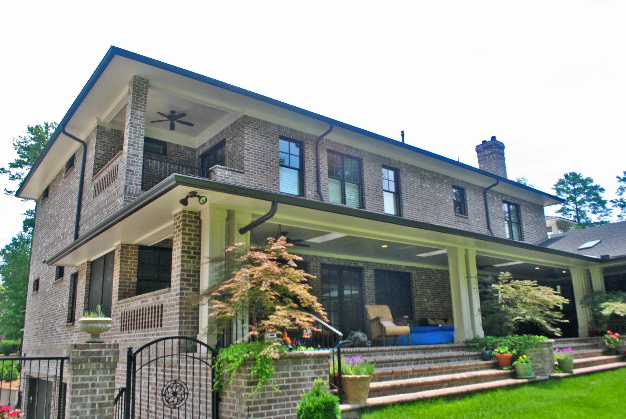 Two-story brick house with a porch, sitting on a landscaped yard with potted plants and trees, under a cloudy sky.