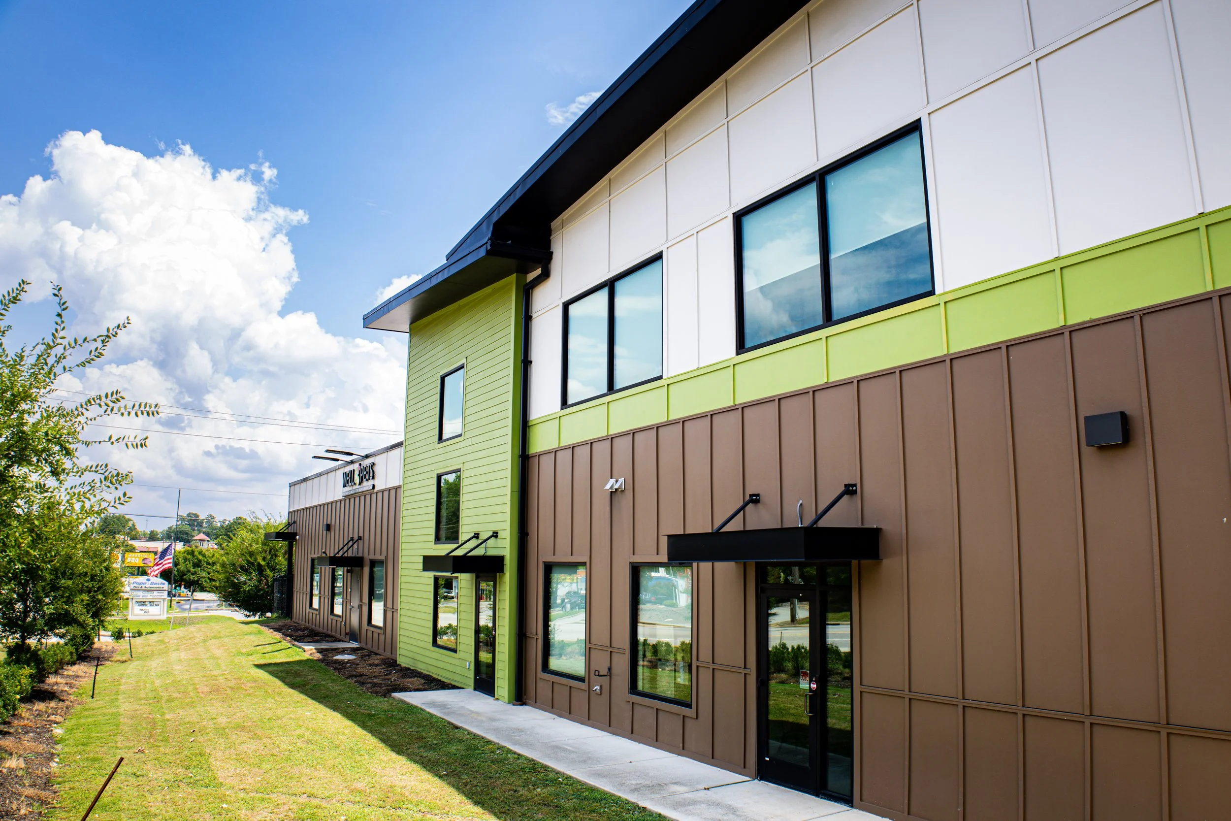 Modern building with colorful exterior panels in green, brown, and white, large windows reflecting the sky, and a well-maintained grassy area with small trees in the foreground under a partly cloudy blue sky.