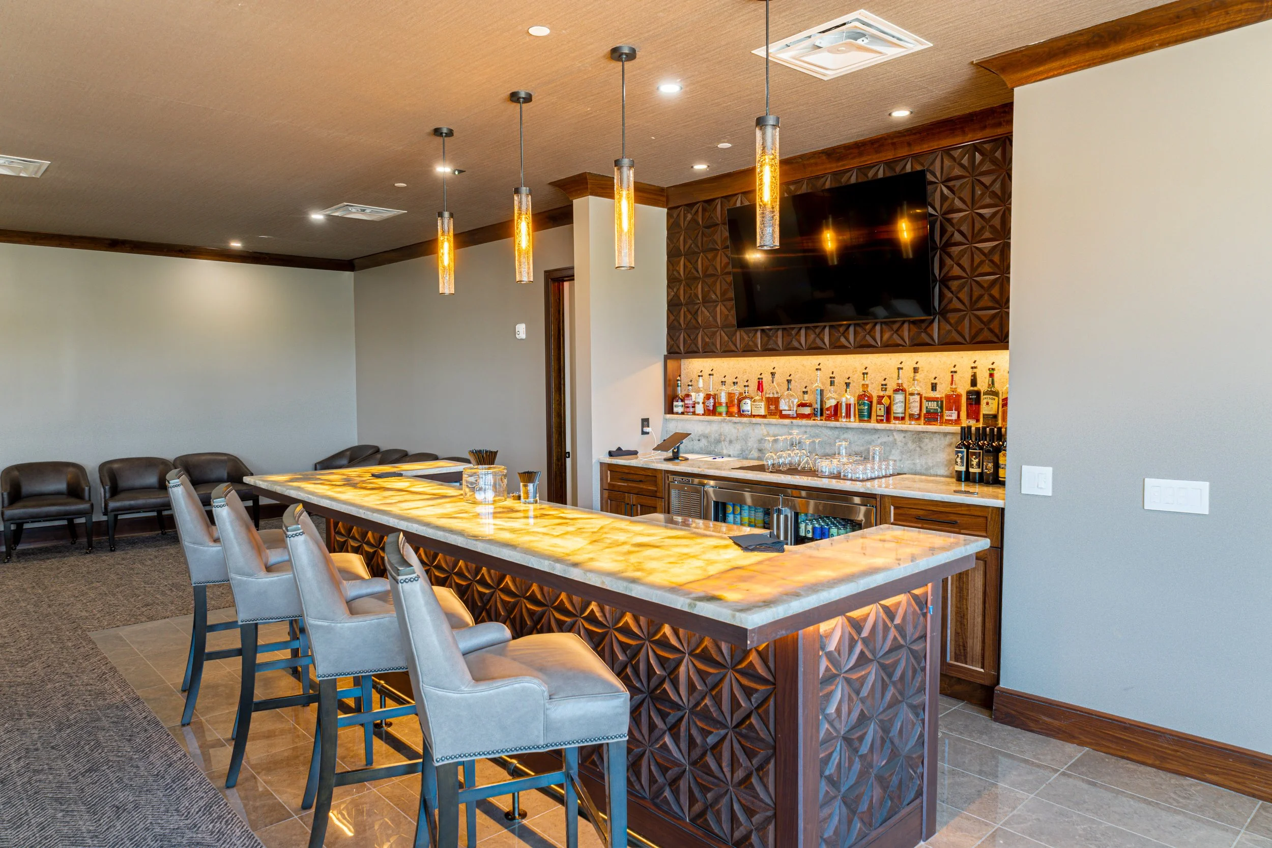 Modern bar area with a marble countertop, several bar stools, hanging pendant lights, a wall-mounted flat-screen TV, and a liquor display behind the bar.