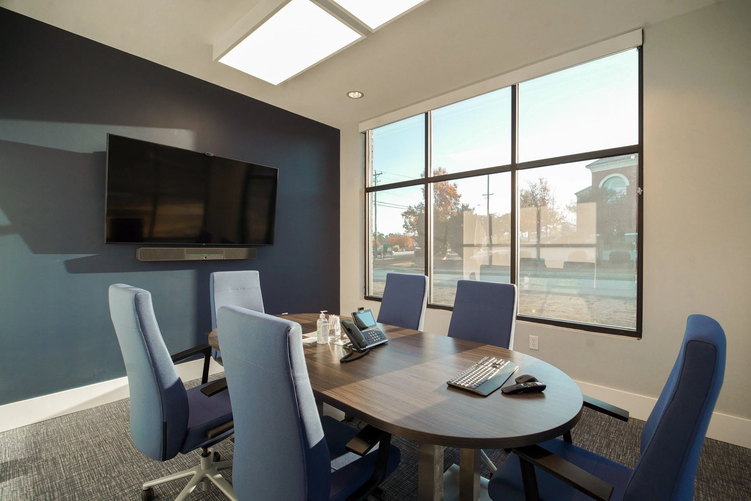 Modern conference room with large windows, a wooden oval table, and blue chairs. A wall-mounted flat-screen TV and a conference phone are on the table, with hand sanitizer and remote controls visible. Bright sunlight streams in through the windows, r