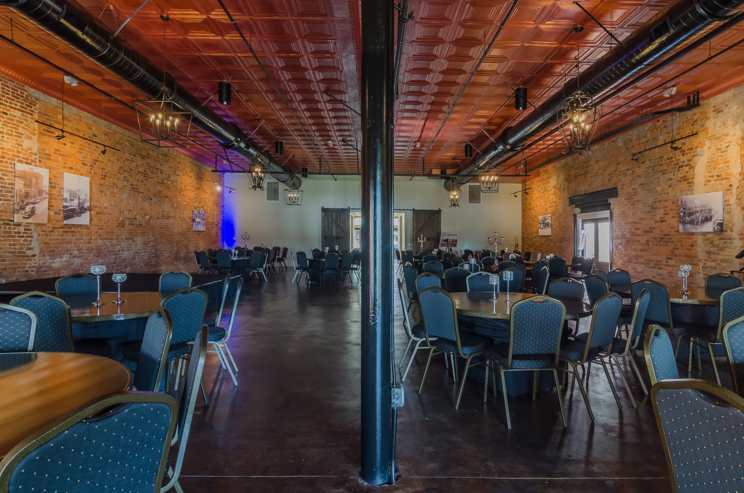 A large room with brick walls, a polished wooden ceiling with black ductwork, several dining tables with blue chairs, and decorative light fixtures hanging from the ceiling.