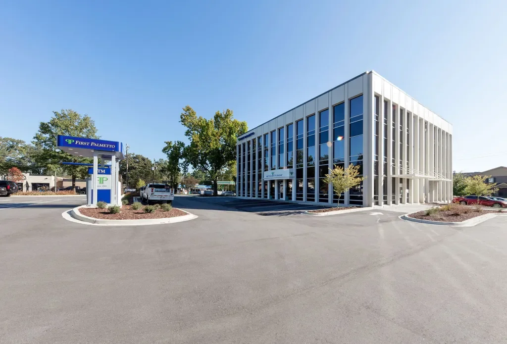 Exterior view of a modern bank building with large glass windows, parking lot in front, and a sign for First Palmetto Bank.