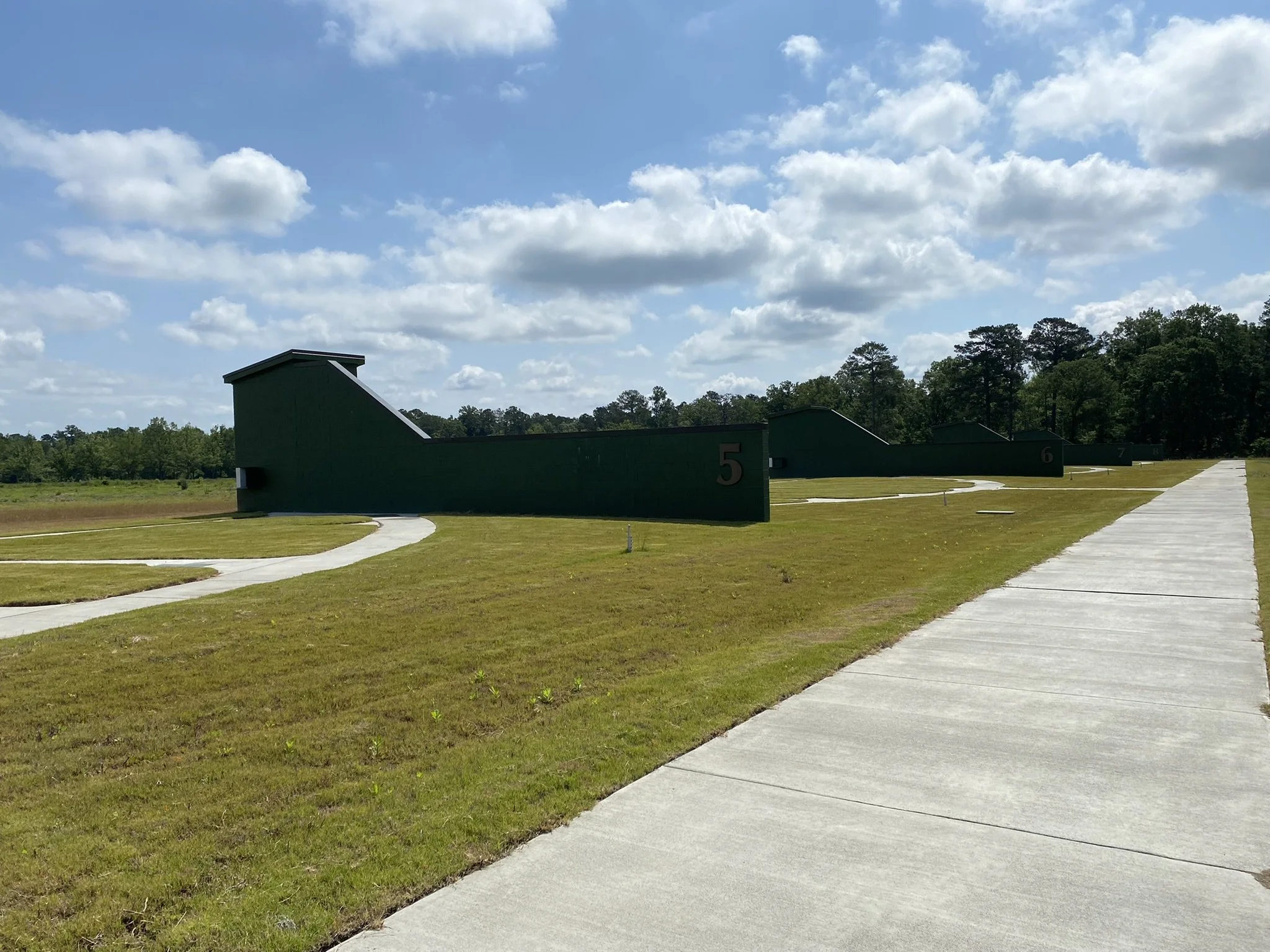Seven green shooting range shelters with numbered signs from 5 to 8, set in an open grassy field with a wooded area in the background, under a partly cloudy sky.