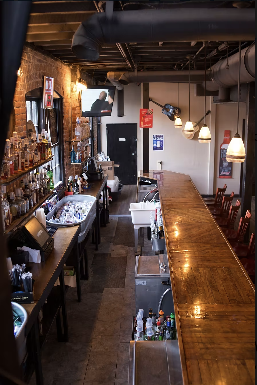 Interior of a bar with a wooden counter, various bottles on shelves behind it, and hanging pendant lights. A TV is mounted on the wall, and there are chairs along the counter.