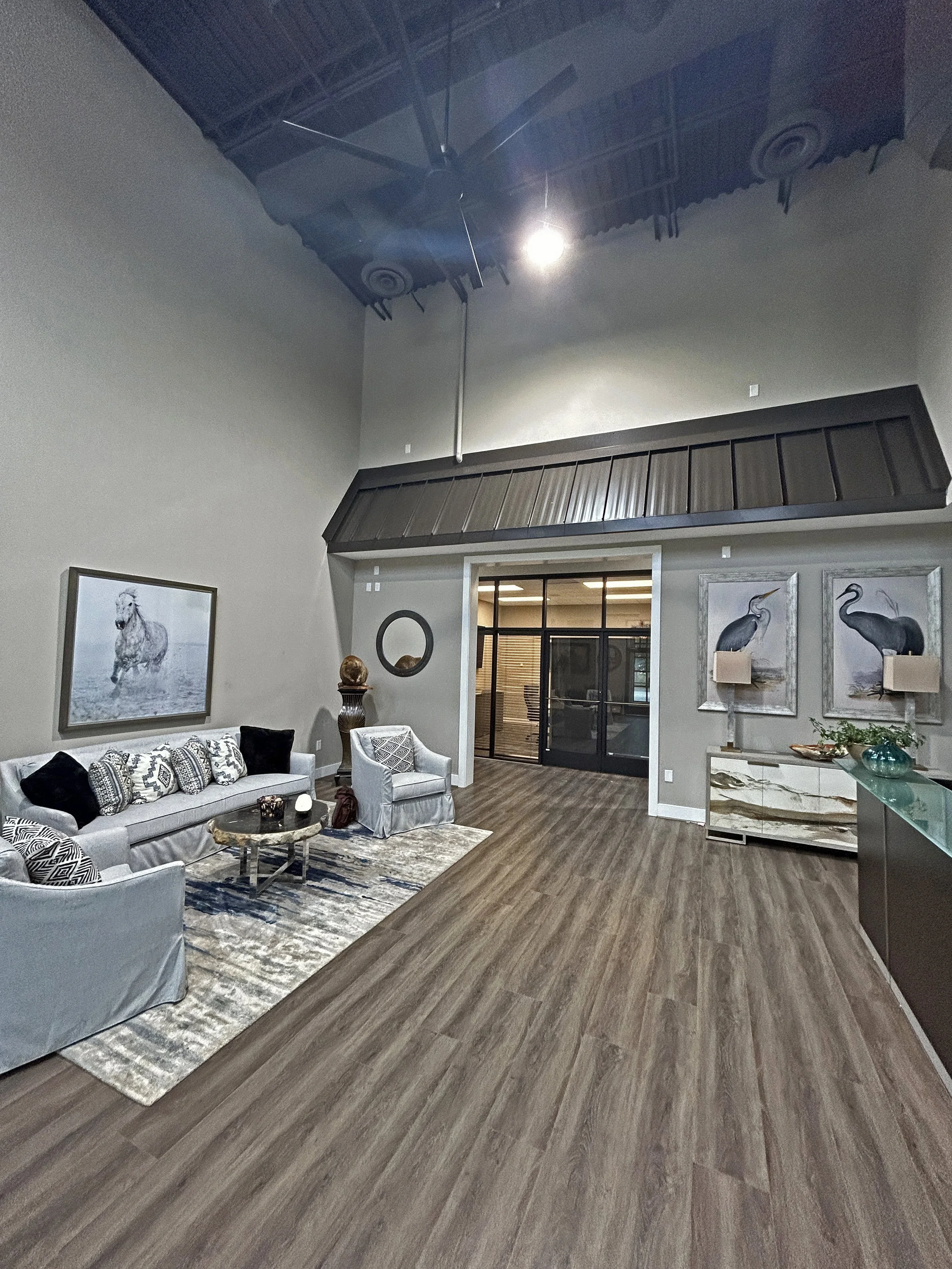 Living room with light-colored sofa, armchair, patterned pillows, modern coffee table, area rug, and wall art featuring animals, next to a sliding glass door with a view of a room with office furniture and decor.