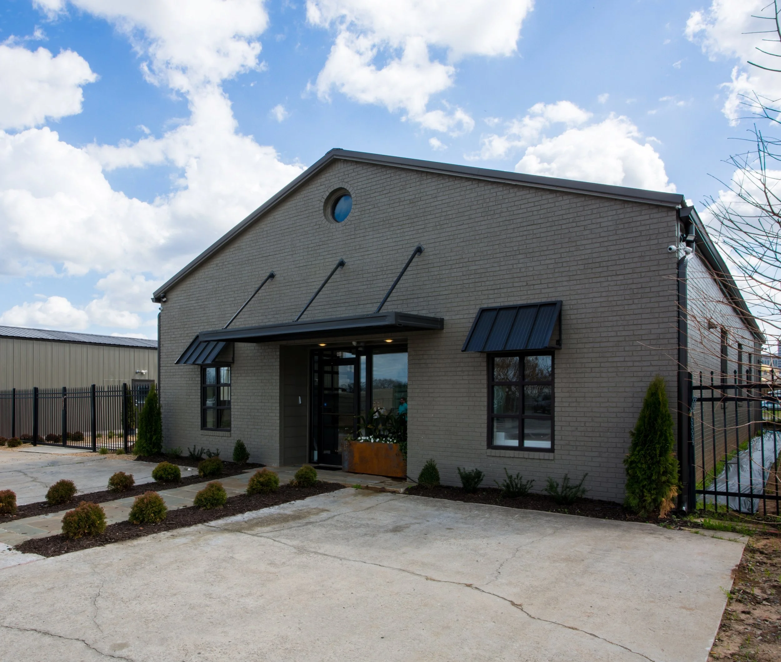 A modern two-story house built with light-colored bricks, featuring black window frames and a sloped roof. The front yard has a concrete driveway with small shrubs planted along the border, and the sky is partly cloudy with white clouds and blue sky.