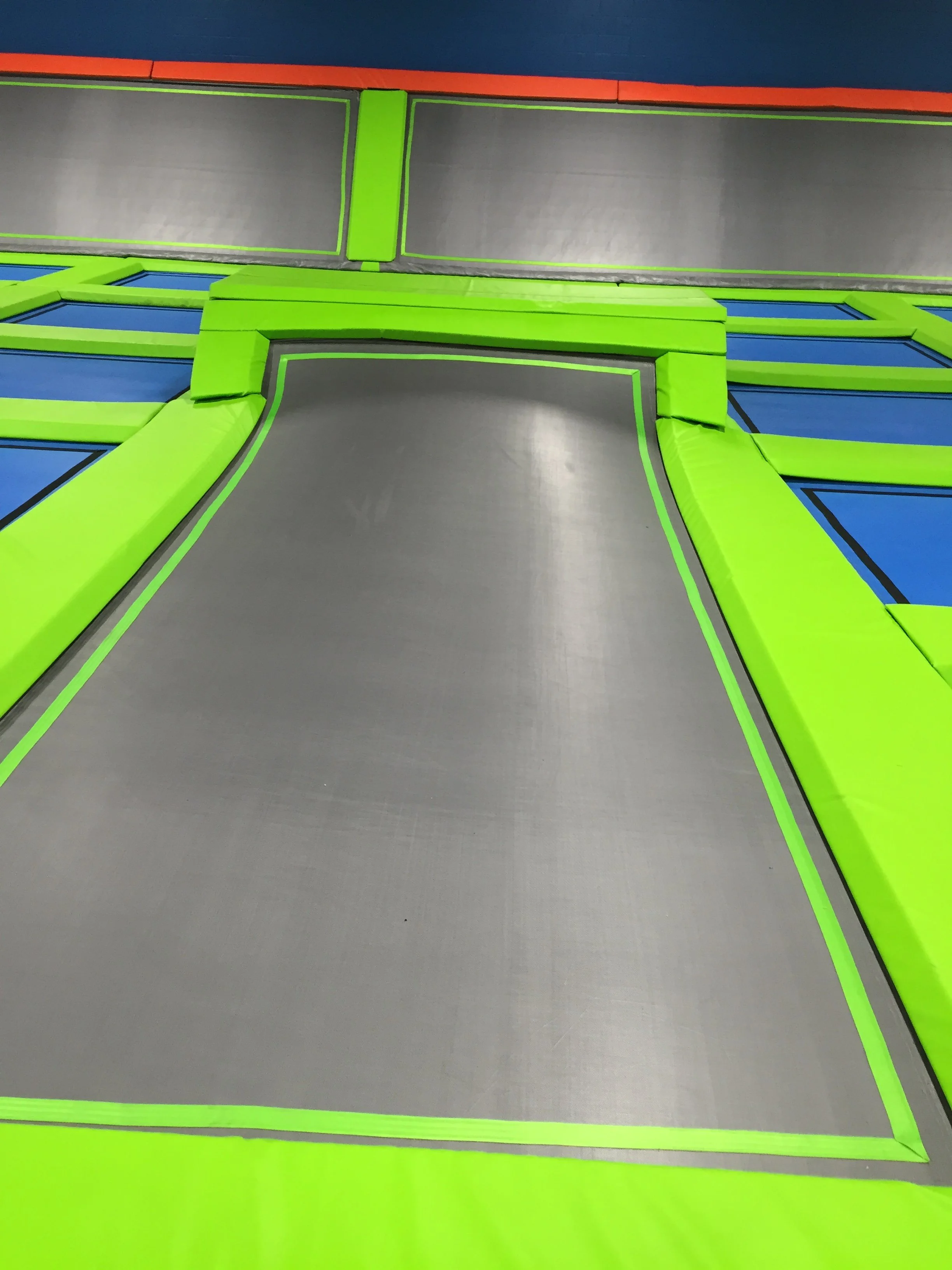 View of an indoor trampoline park with multiple interconnected trampolines, including a large rectangular one in the center surrounded by padded green and blue areas for safety.