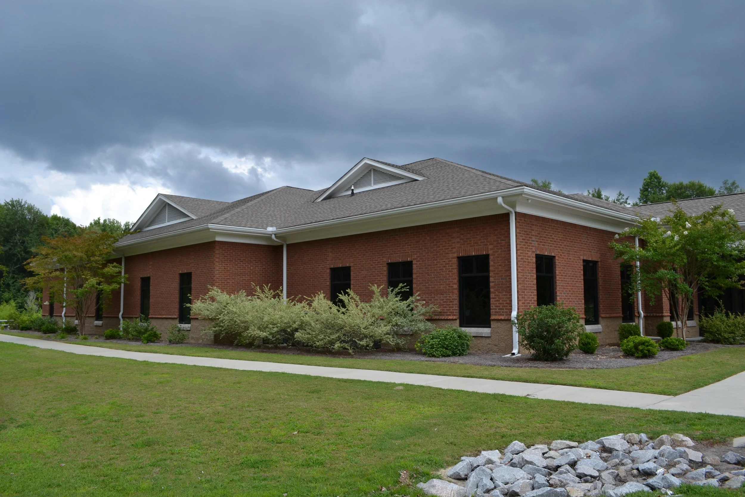 A brick building with a gray shingle roof and black windows, surrounded by green trees, shrubs, and a well-maintained lawn, with a cloudy sky overhead.