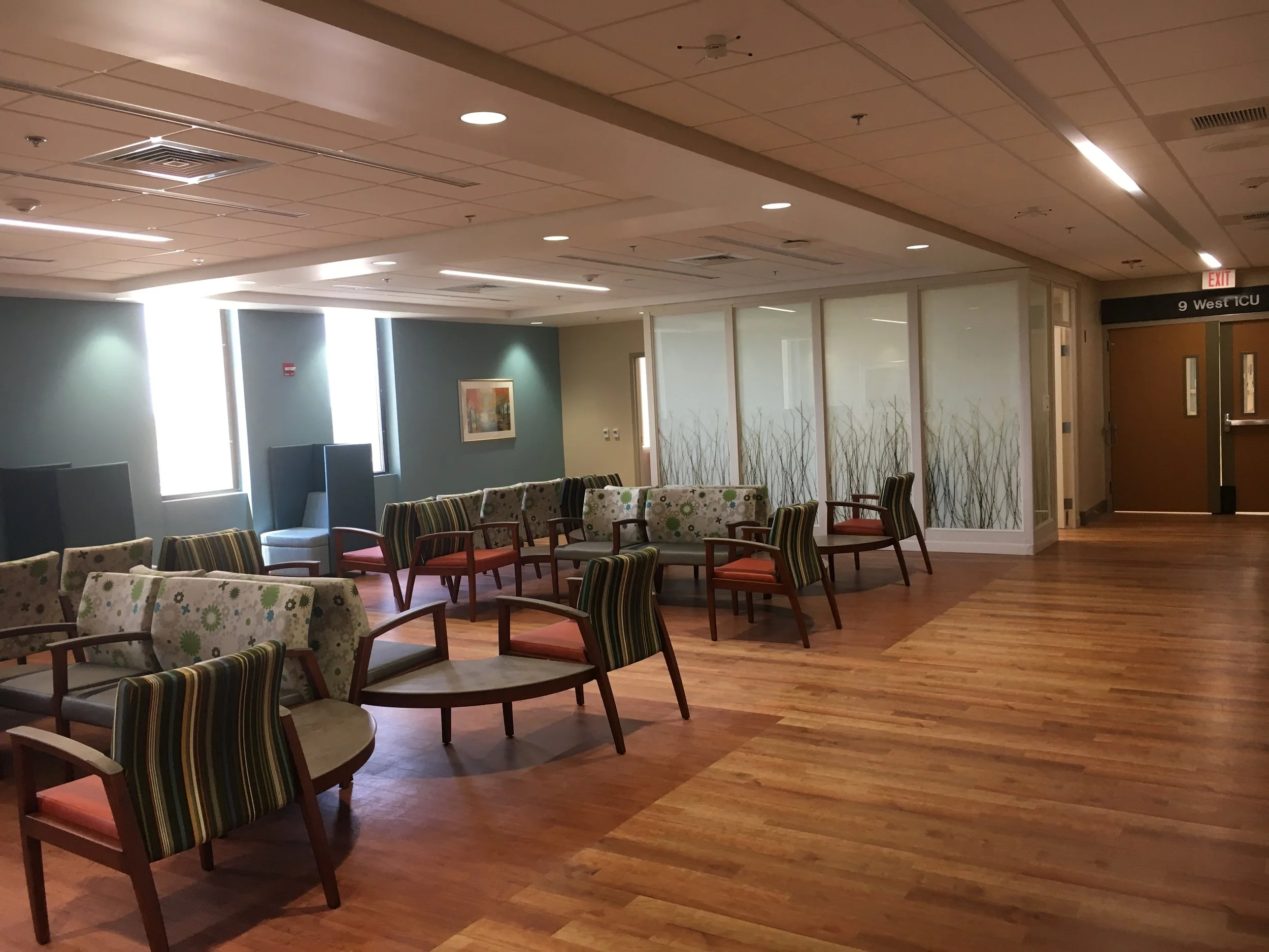 Empty waiting room with chairs and sofas, wood flooring, large windows, and a sign indicating 