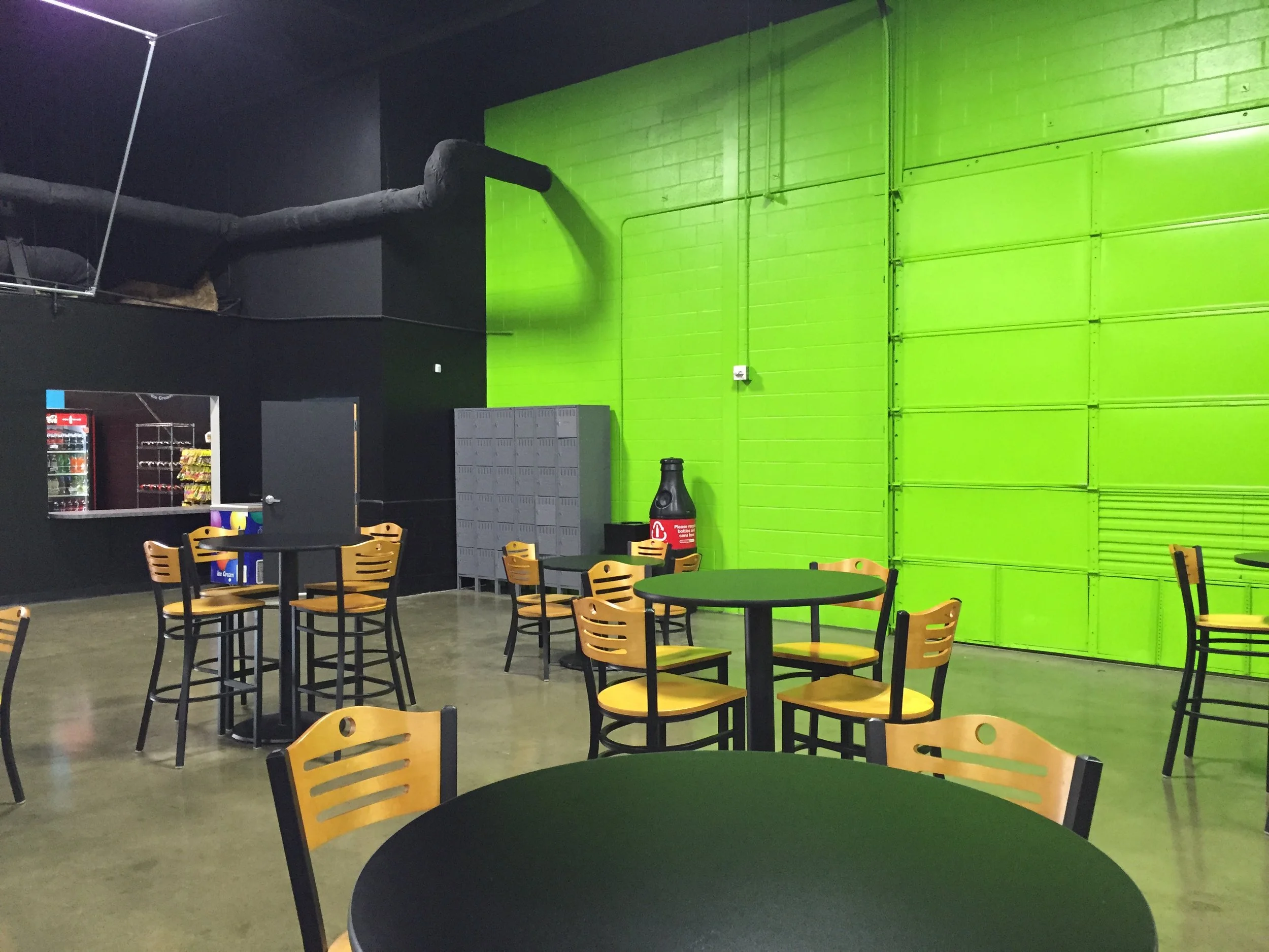 Interior of a cafeteria or lounge area with black tables and yellow chairs, green wall, lockers, and a beverage vending machine in the background.