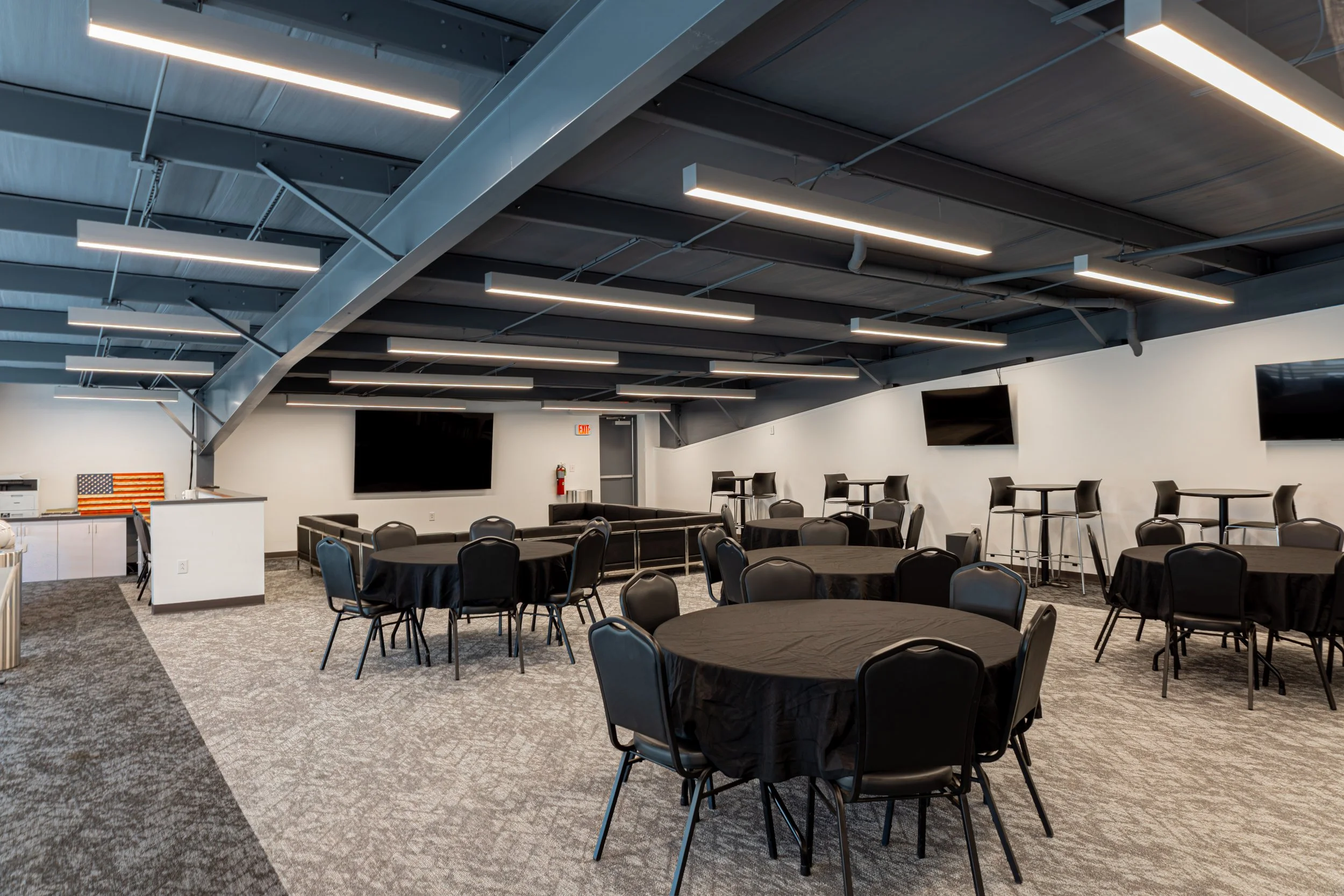 An indoor event space with round tables covered with black tablecloths and black chairs. The ceiling has modern linear lighting and exposed ductwork, with a large flat-screen TV on the wall. There is a kitchenette area with an American flag decoration and a fire extinguisher nearby.