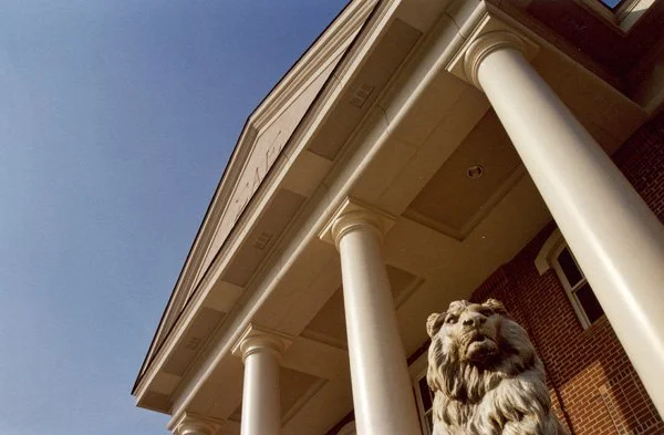 A close-up of a building with large white columns and a lion statue at the base of one column, under a clear blue sky.
