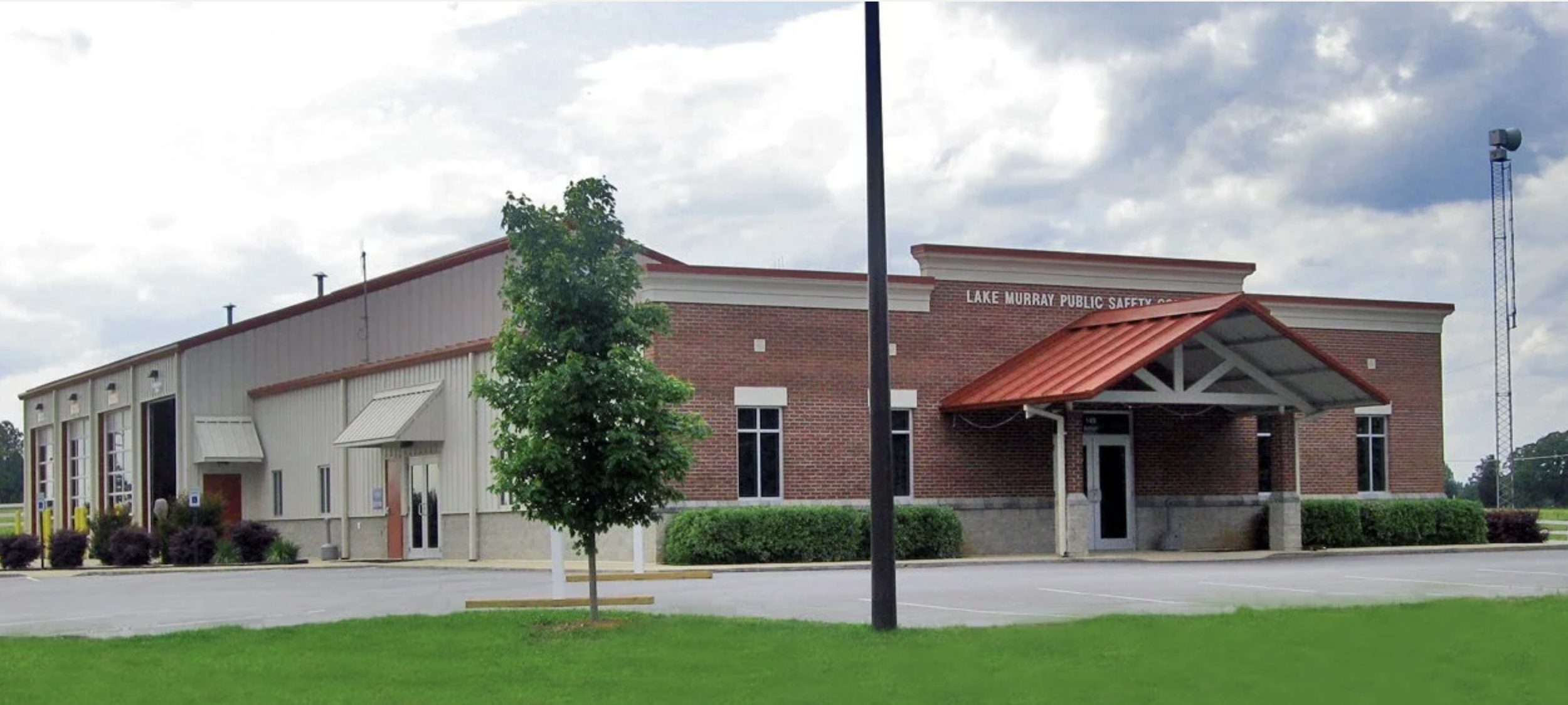 A building with a sign that reads 'Lake Murray Public Safety'. It has a combination of brick and metal siding, with a red metal roof over the entrance. There is a small tree and a parking lot in front of the building, with some shrubs and grass nearby. The sky is cloudy.