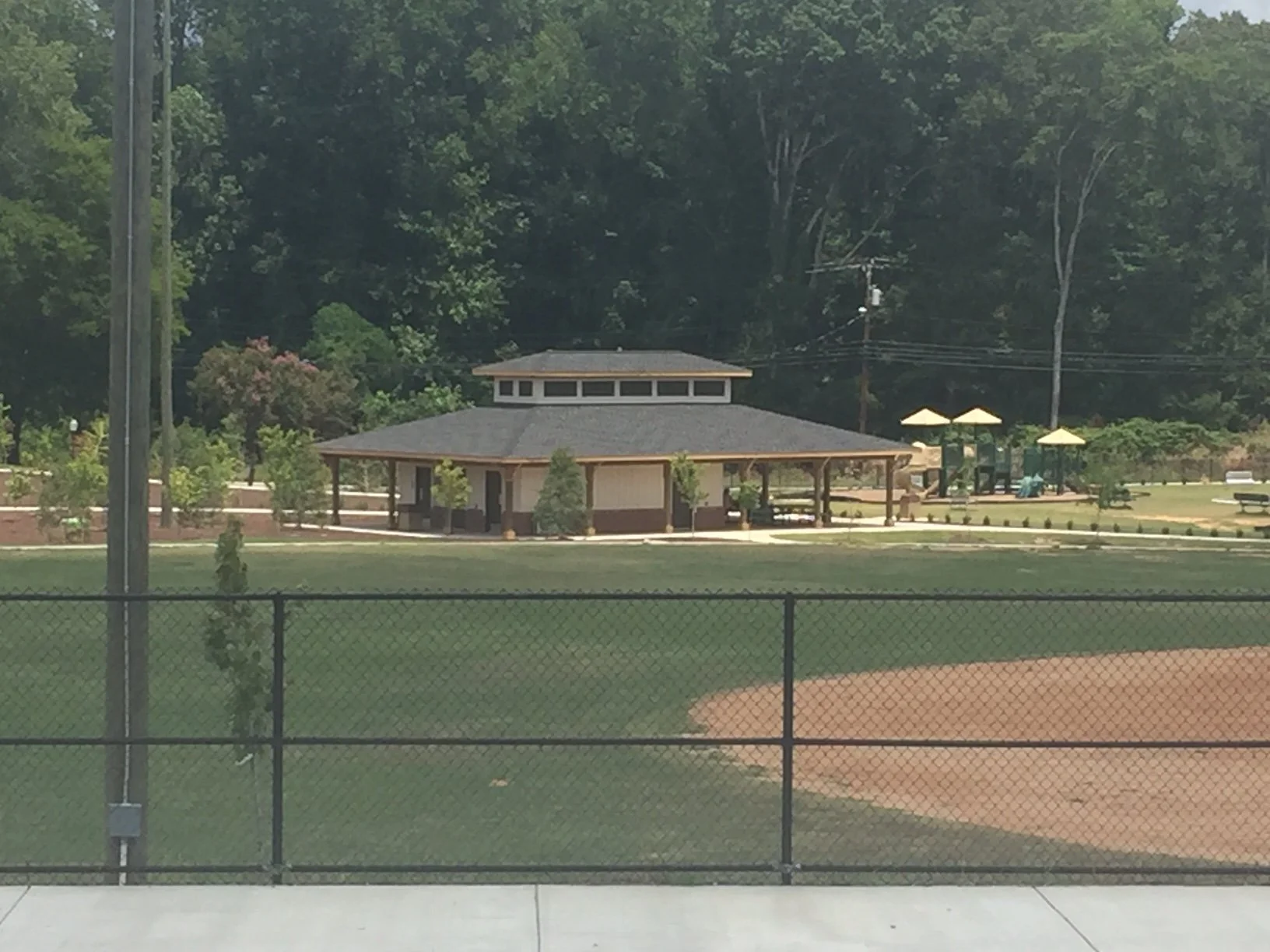 A baseball field with a fence in the foreground, a small building with a roof, a playground with shaded structures, and trees in the background.