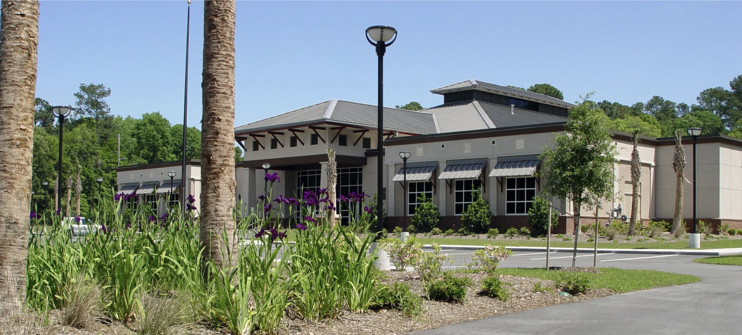 A modern building with large windows, surrounded by landscaped greenery, trees, and purple flowers, under a clear blue sky.