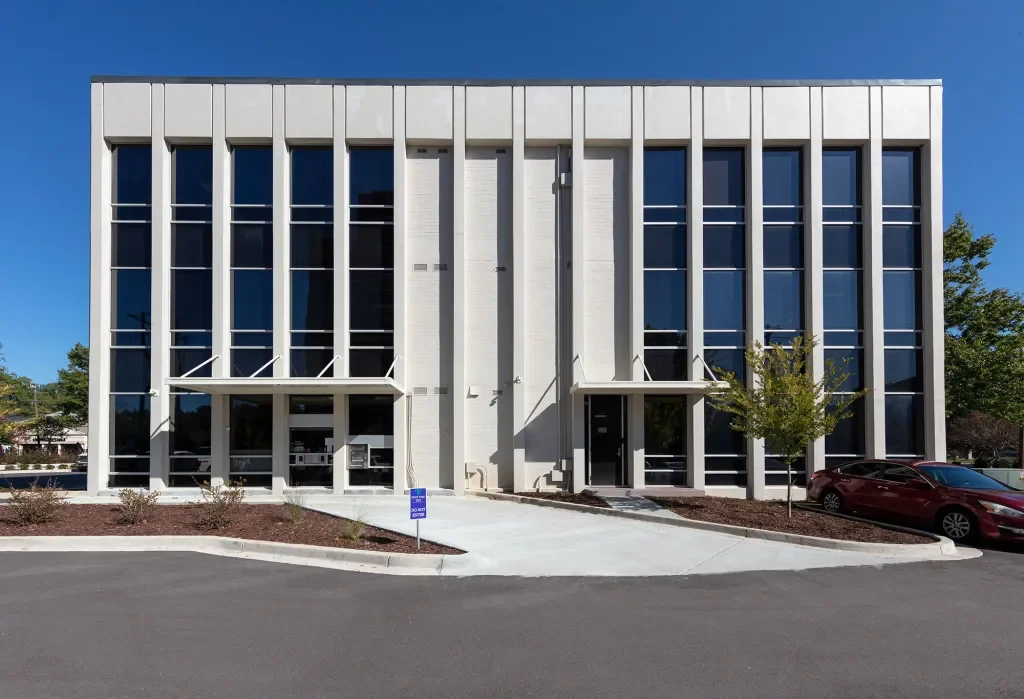 A modern, white, three-story office building with tall vertical windows and a centered entrance, surrounded by a parking lot and landscaped area with small trees and bushes.