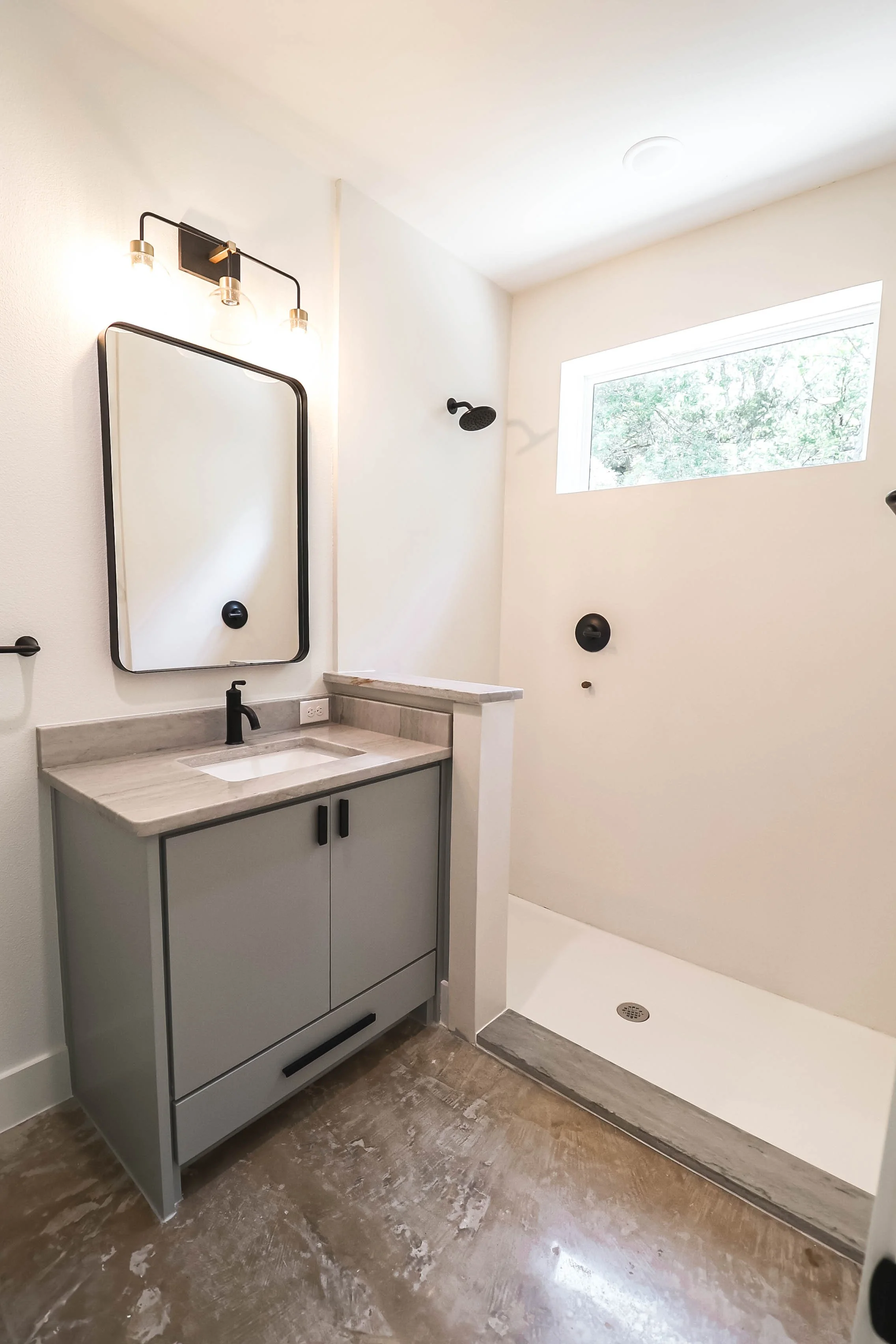 Modern bathroom with gray vanity, marble countertop, wall-mounted black faucet, wall mirror, black light fixture, walk-in shower with black shower head, large horizontal window, white walls, wood-look flooring.