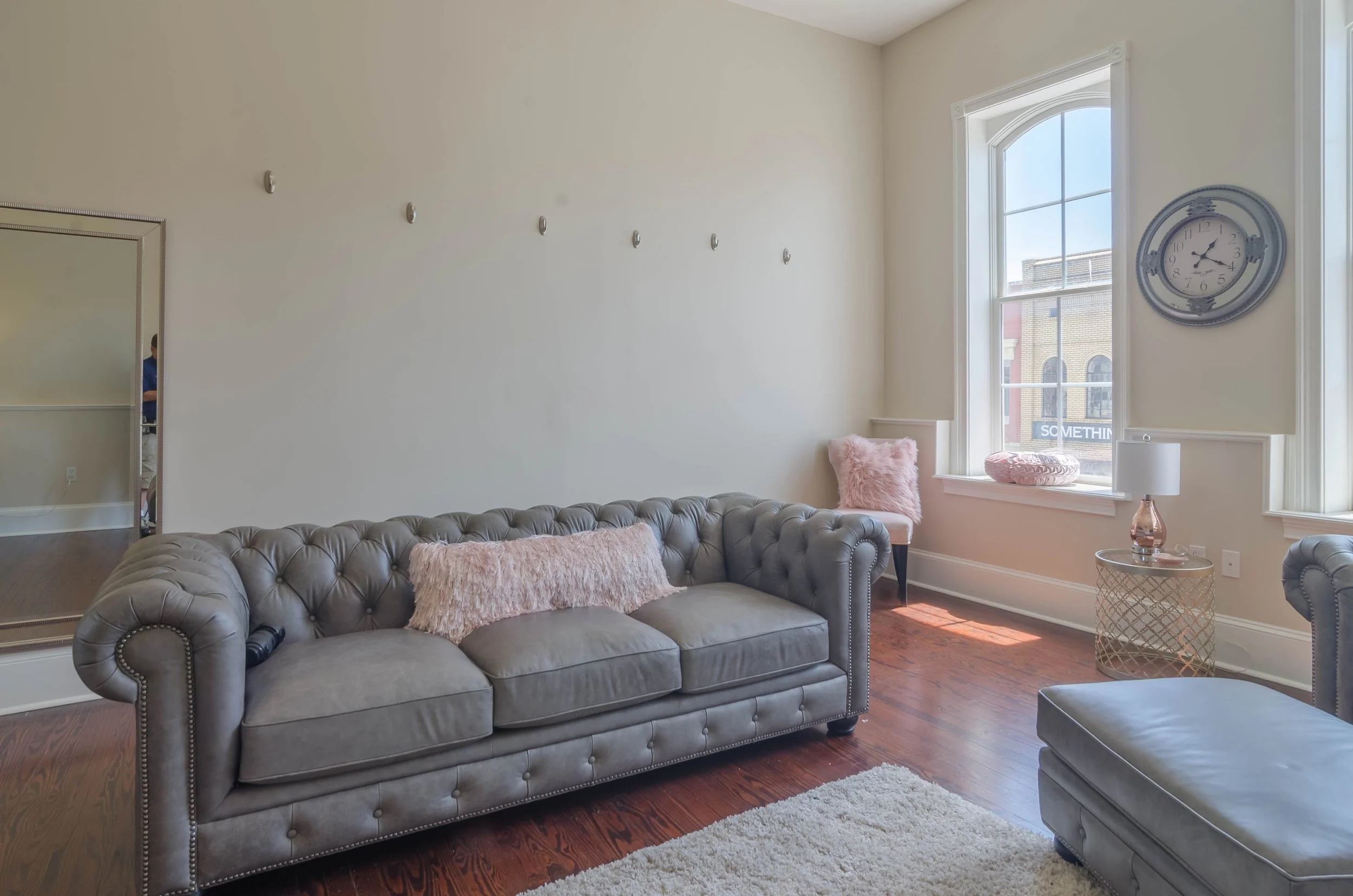 Living room with a tufted gray sofa, a pink fluffy pillow, a small side table with a lamp, large window with a view of brick buildings, a round wall clock, a mirror, and hardwood floors.