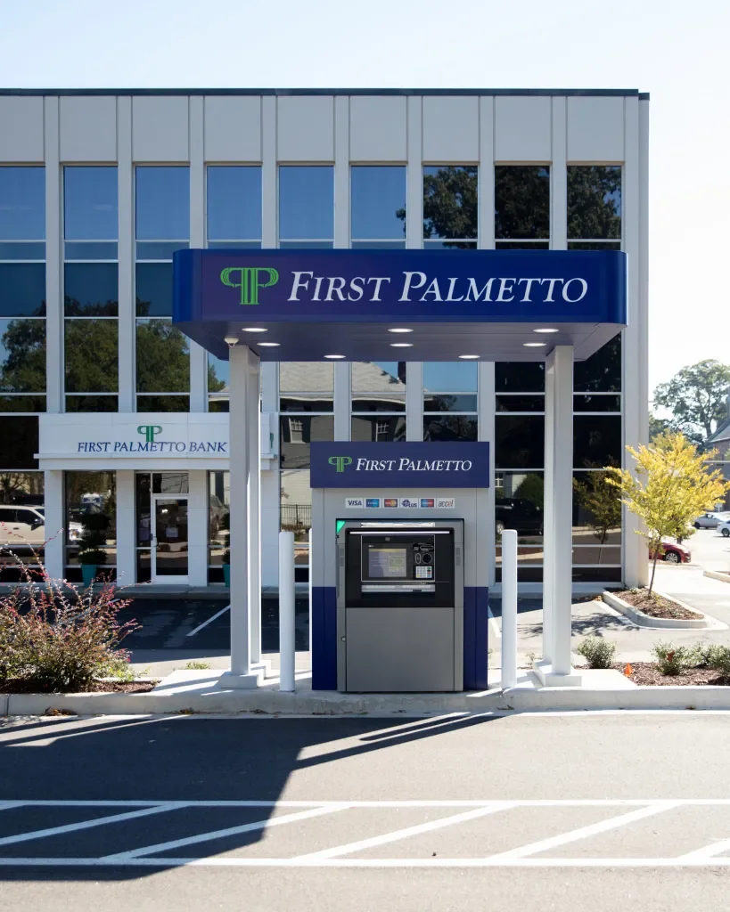ATM kiosk in front of First Palmetto Bank building with outdoor signage, parking lot, and landscaped area with small plants and trees.