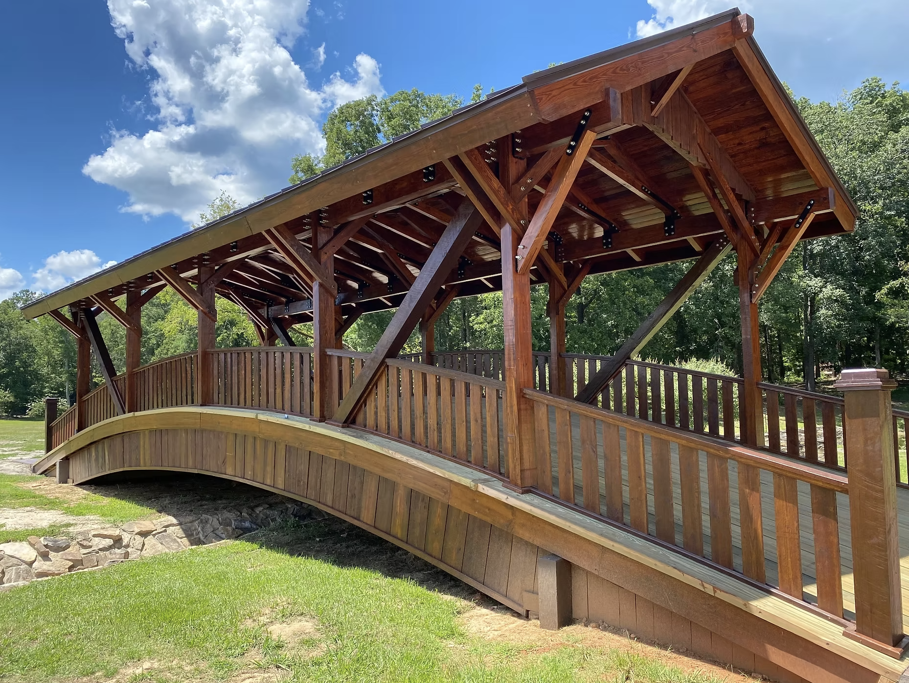 A wooden bridge with an arched design, railings, and a gabled roof, situated in a park-like setting with green trees, grass, and a partly cloudy blue sky.