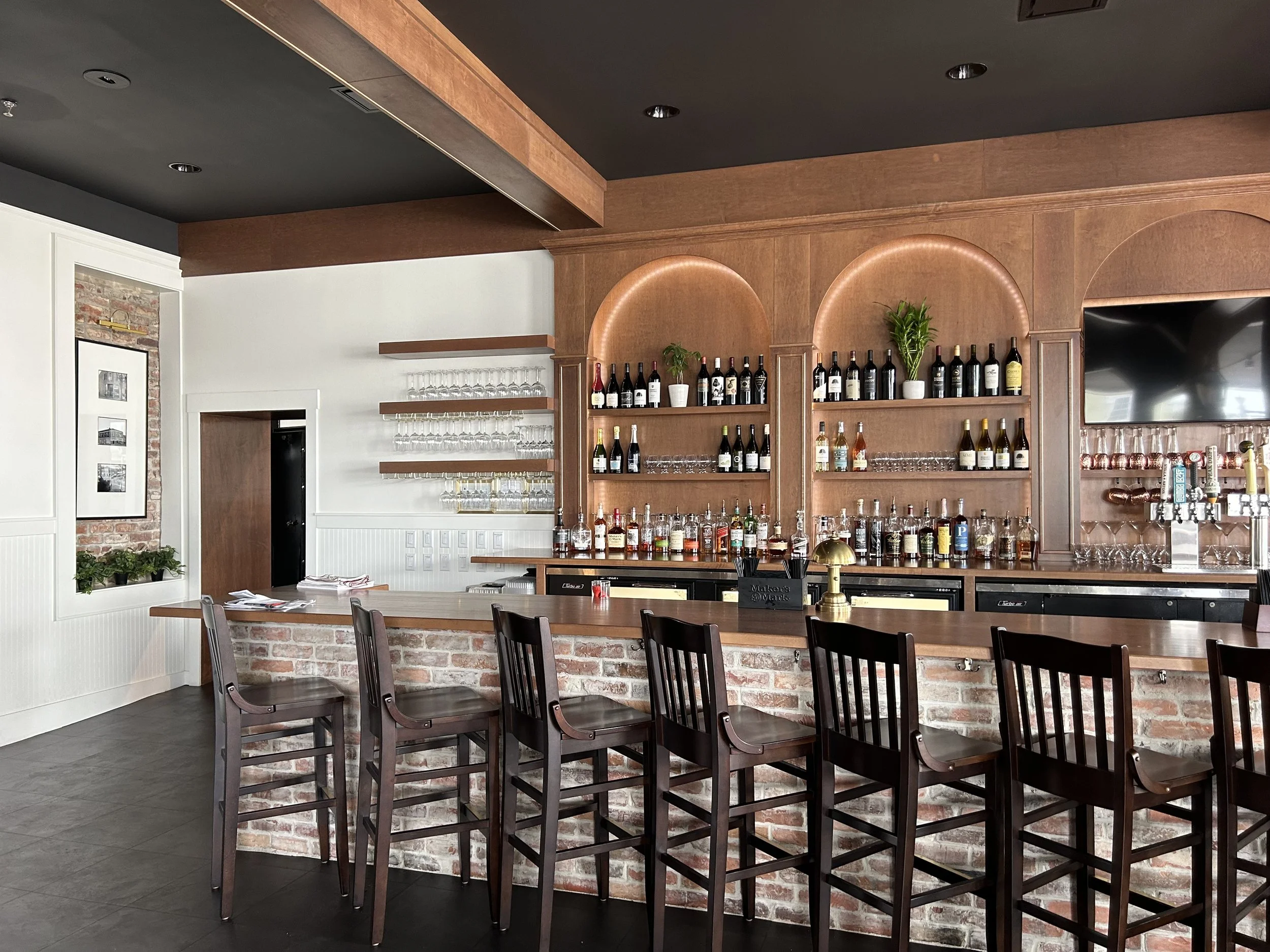 Interior of a restaurant or bar with a wooden bar counter, alcohol bottles on shelves, and multiple wooden chairs around the bar.