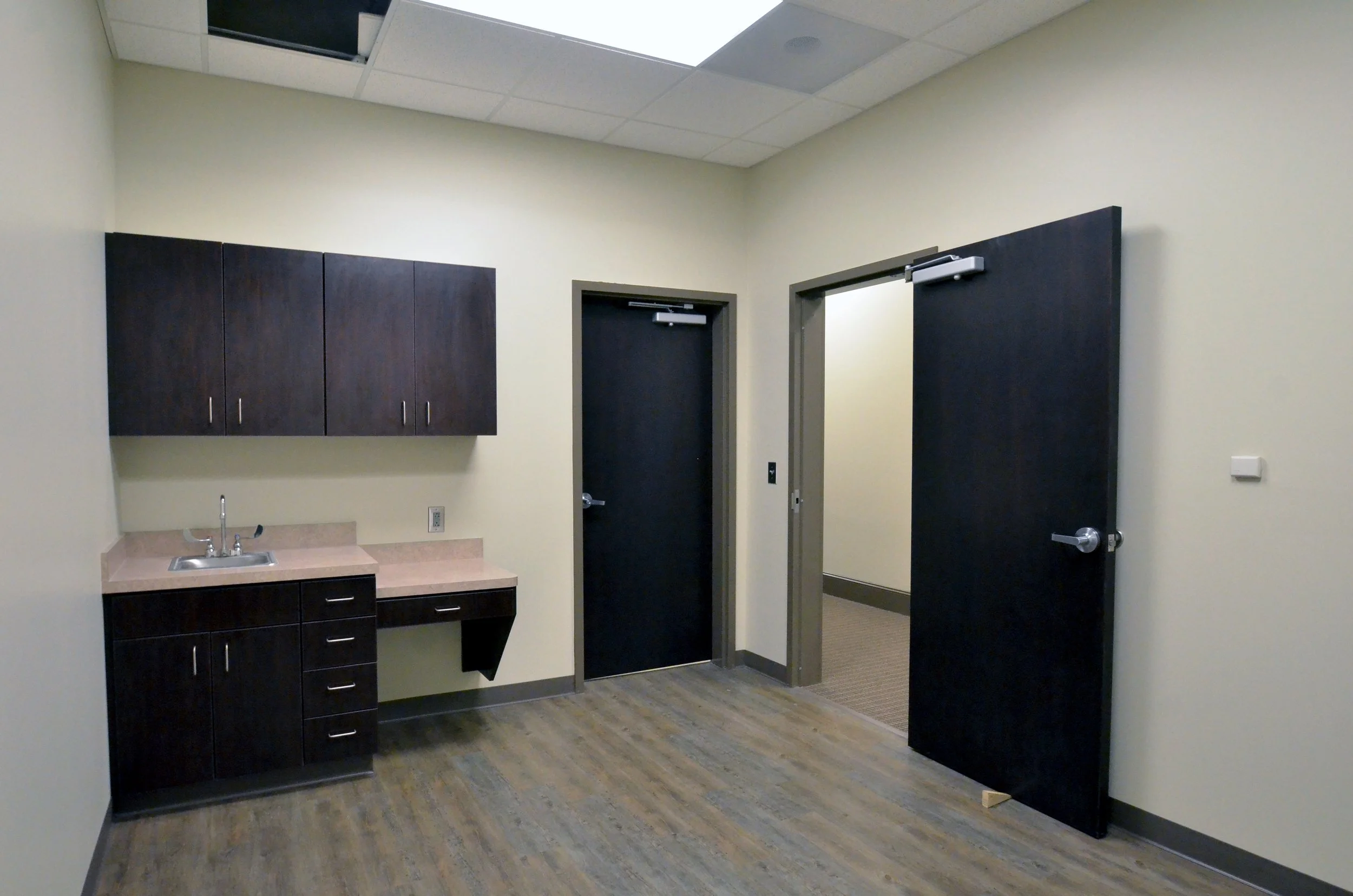 An empty room with a small kitchen area, including dark cabinets, a sink, and a mirror, with two closed dark doors and a beige wall.