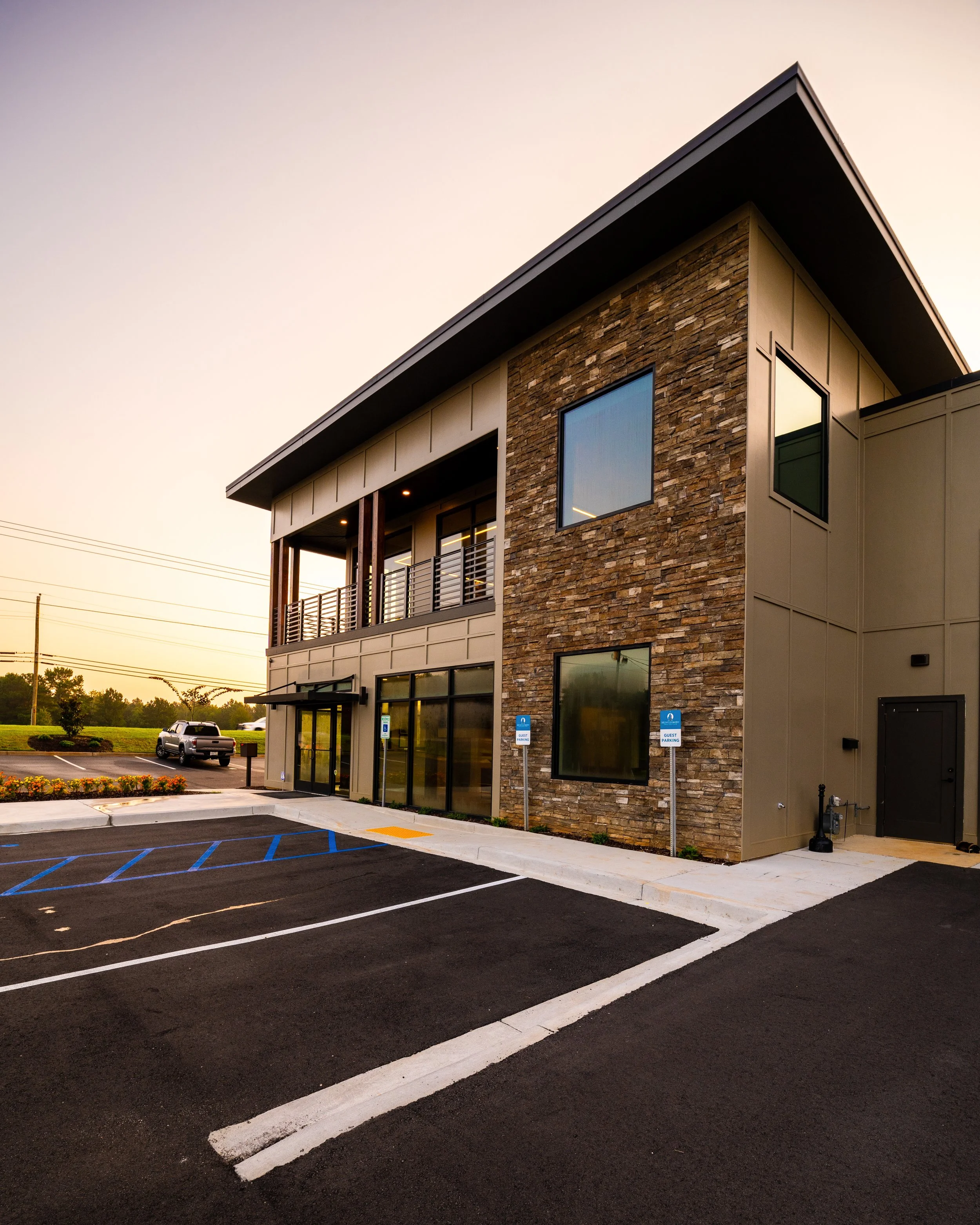 Modern commercial building with stone and beige siding, large windows, and a balcony, under a clear evening sky, with a parking lot in front marked for handicapped parking.