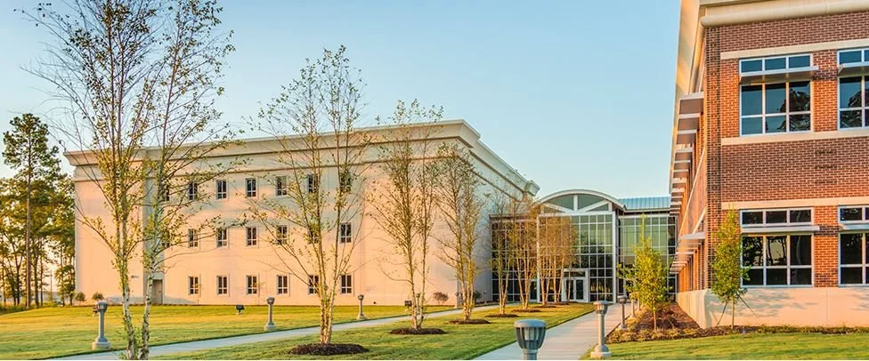 Campus with modern white and brick buildings, green lawn, trees, and walkway.
