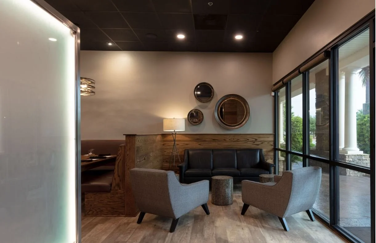 A modern indoor lounge area with a black leather sofa, two light gray armchairs, a floor lamp, and three round wall mirrors, with large windows on the right side allowing natural light.
