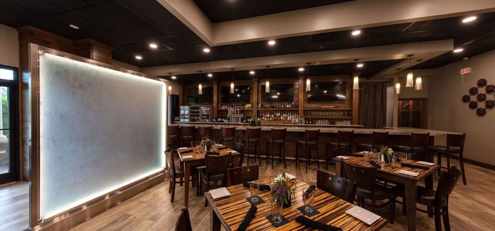 Empty interior of a restaurant or bar with tables set with black napkins, glasses, and flowers, a bar with stools in the background, and warm lighting.