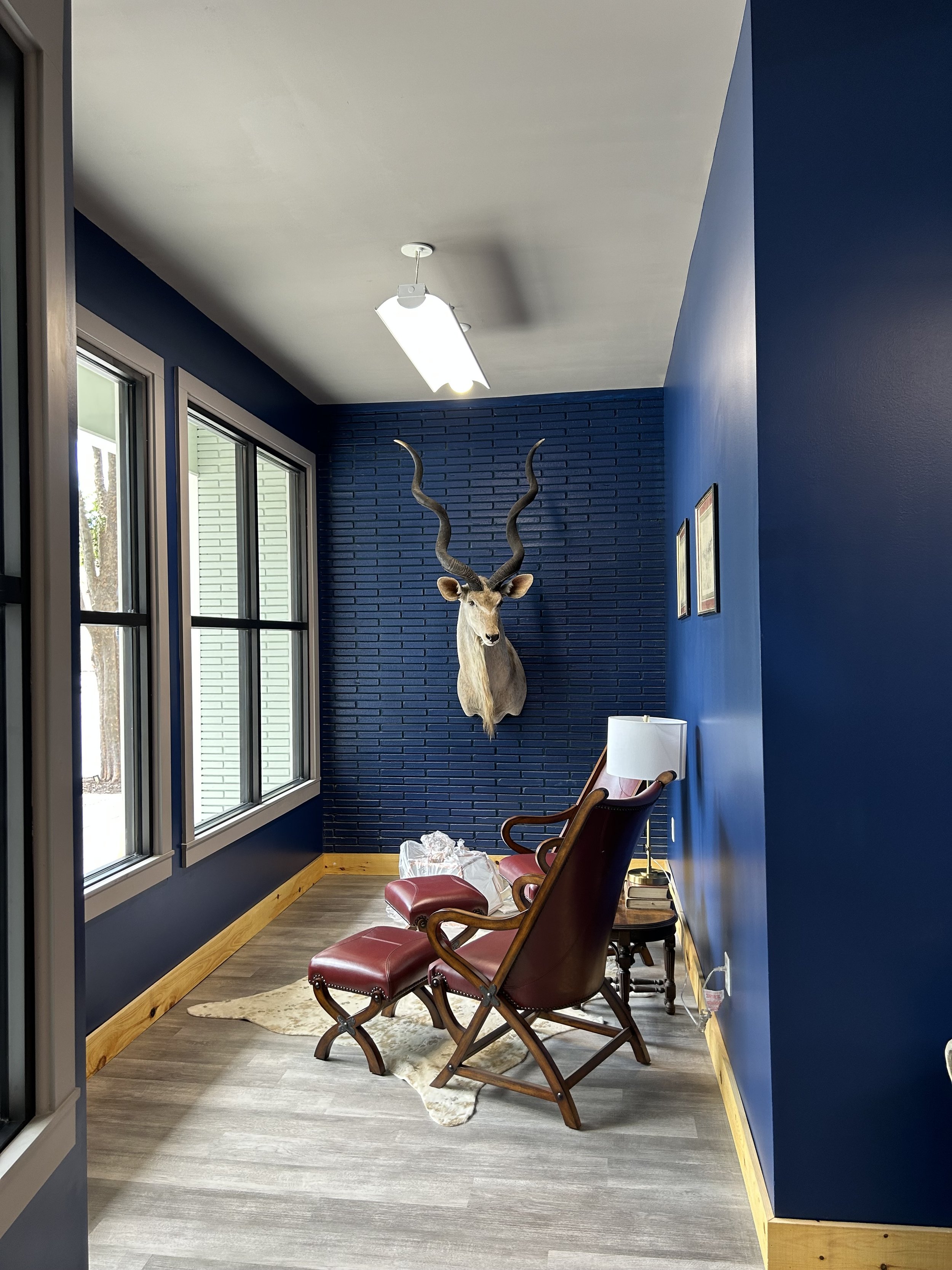 Room with blue walls, three wooden chairs with red cushions, a floor lamp, a mounted deer head on the dark blue brick wall, three framed pictures, and windows on the left side letting in natural light.