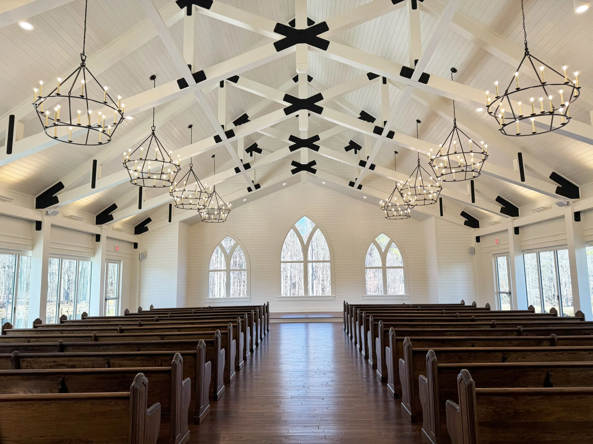 Interior of a church with white wooden walls and ceiling, large arched windows, wooden pews, and multiple black chandeliers with candle-like lights hanging from the ceiling.