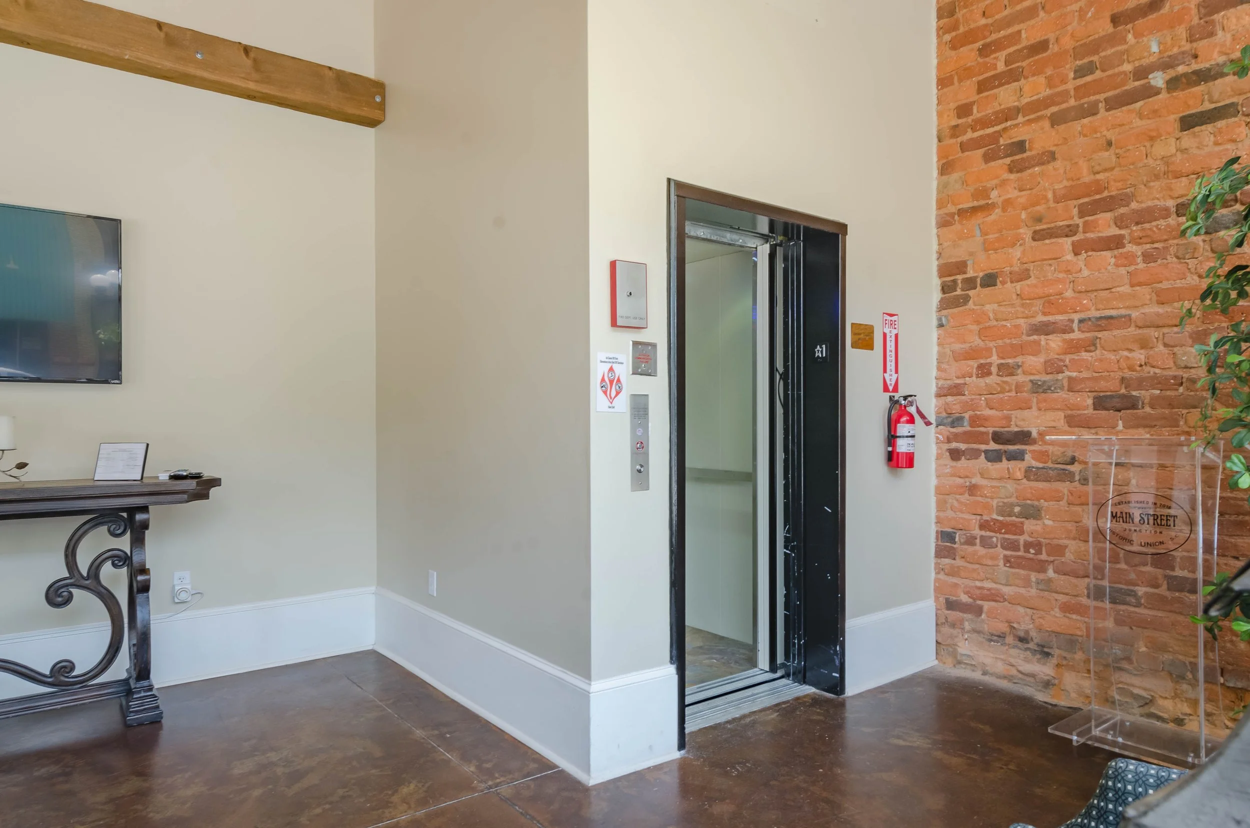 Interior of a building lobby featuring an open elevator with black frame, exposed red brick wall on the right, a wall with a fire extinguisher and safety signs, a brown floor, and contemporary decor including a decorative table and a potted plant.