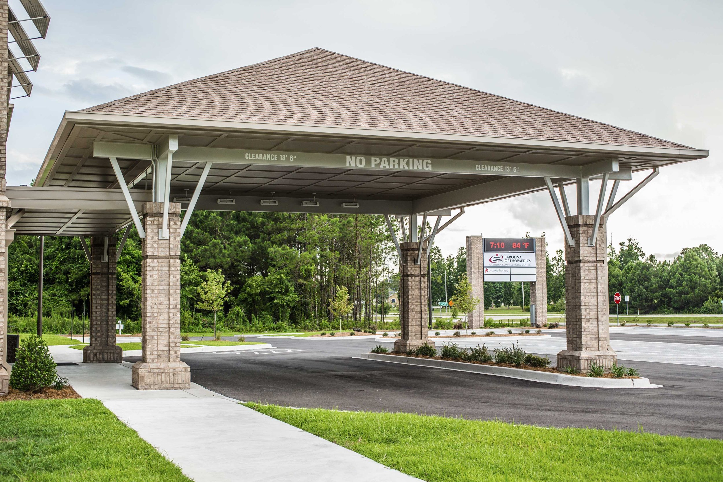 A covered parking area with brick columns, a tiled roof, and a digital sign showing the time, temperature, and an advertisement for Carolina Orthopaedics. There is a sidewalk and green grass in the foreground, with trees and a cloudy sky in the backg