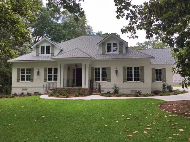 A white two-story house with a front porch, multiple windows with shutters, and a gray shingle roof, surrounded by a neatly maintained lawn and trees.