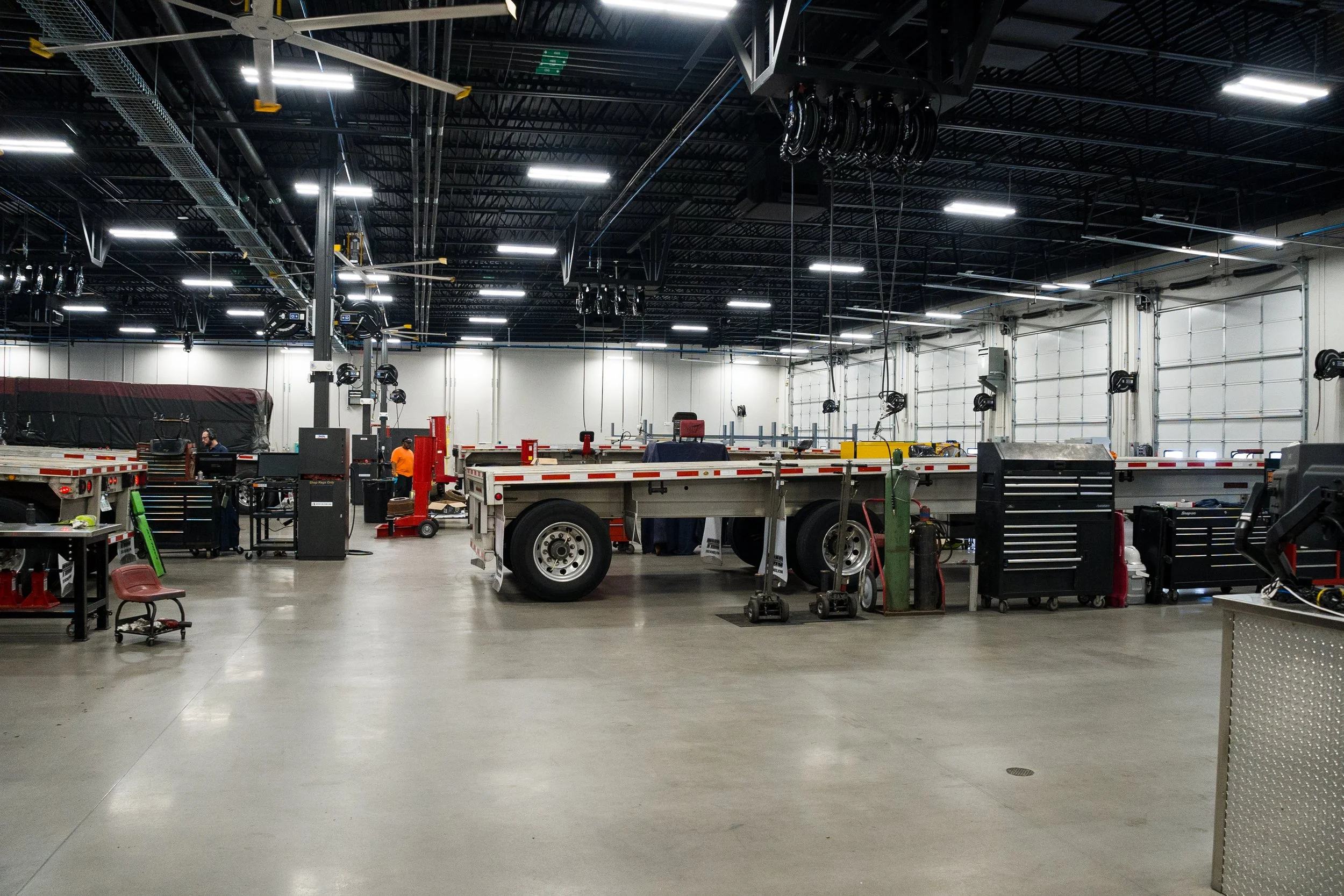 Inside a large warehouse or industrial building with a high black ceiling, bright fluorescent lighting, and a spacious open floor. There are work tables, equipment, and machinery, with a few workers visible in safety gear.