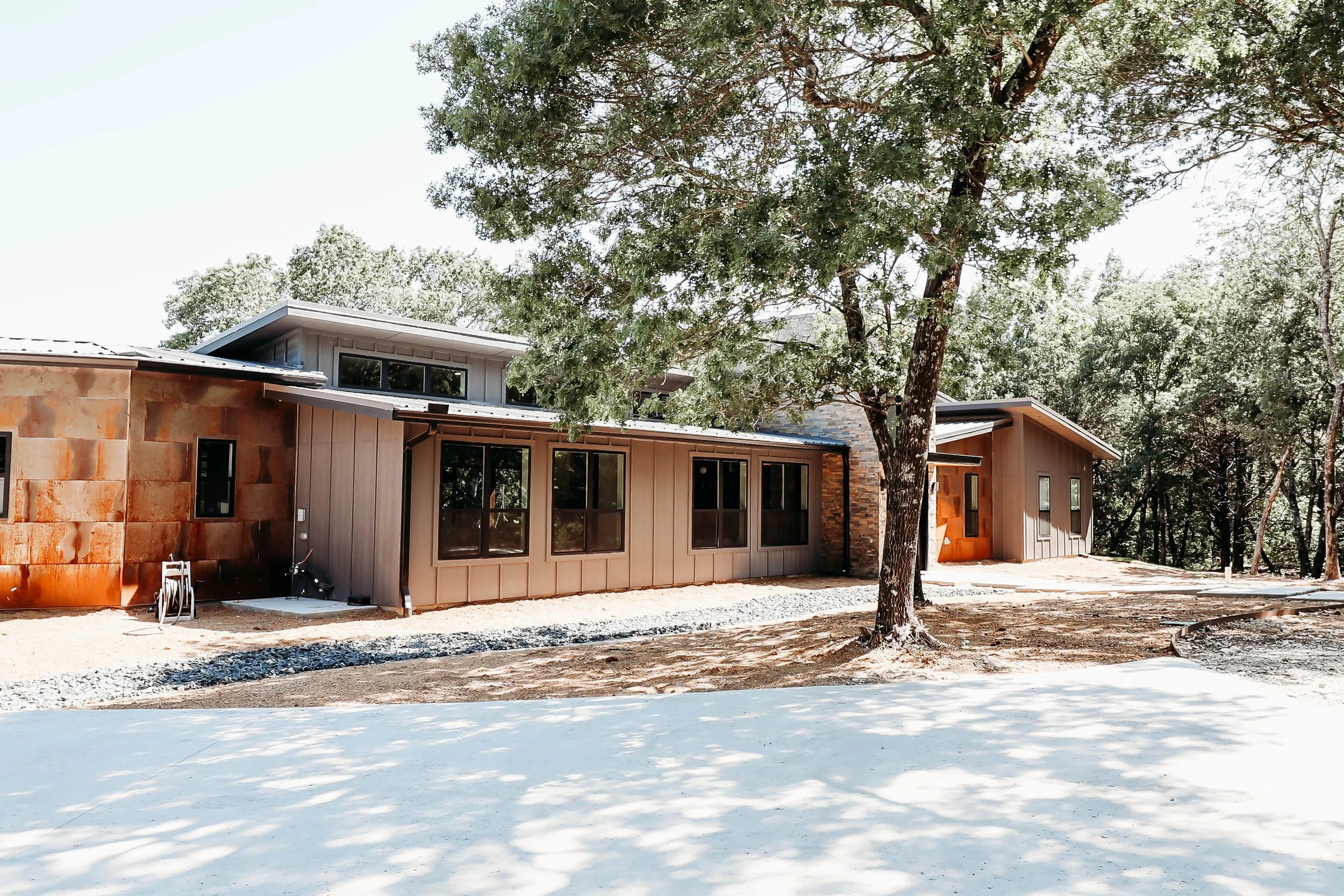 A modern house under construction in a wooded area, with unfinished brick and siding walls, large windows, and a gravel pathway in front, surrounded by trees and a dirt yard.