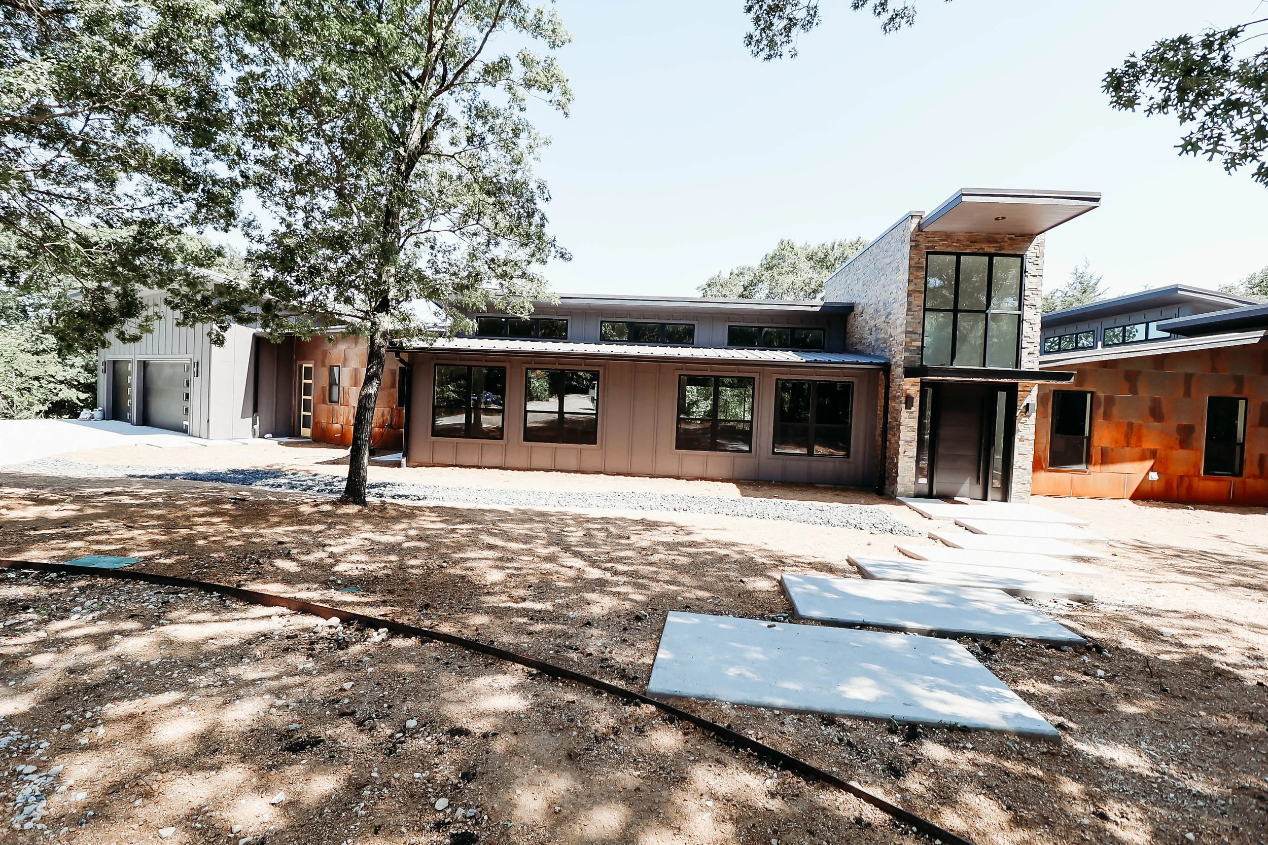 Modern house under construction with large windows, brick accents, and a metal roof in a wooded area.