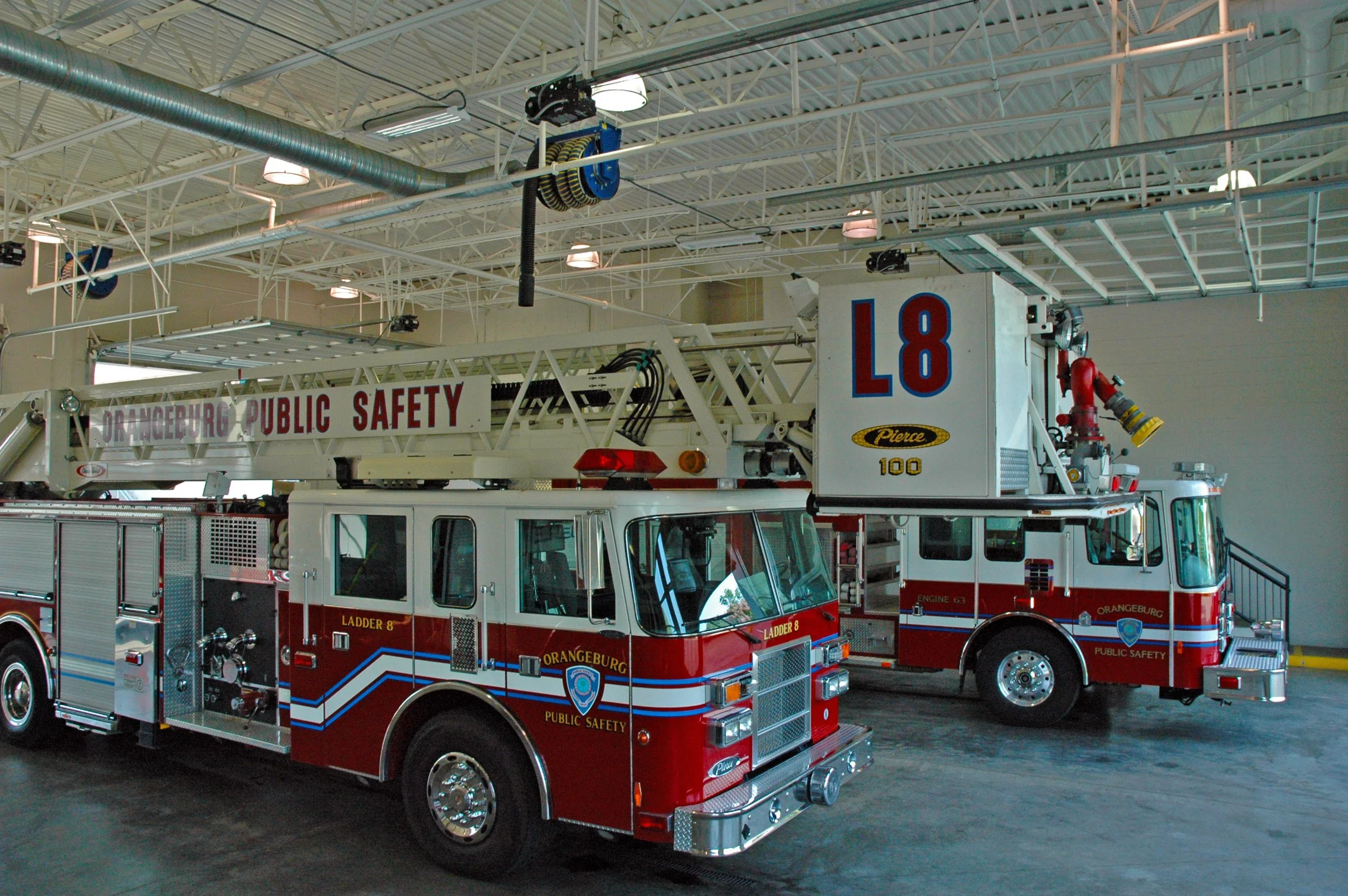 Fire trucks inside a fire station, labeled Orangeburg Public Safety, with ladders and equipment, parked indoors under a high ceiling.