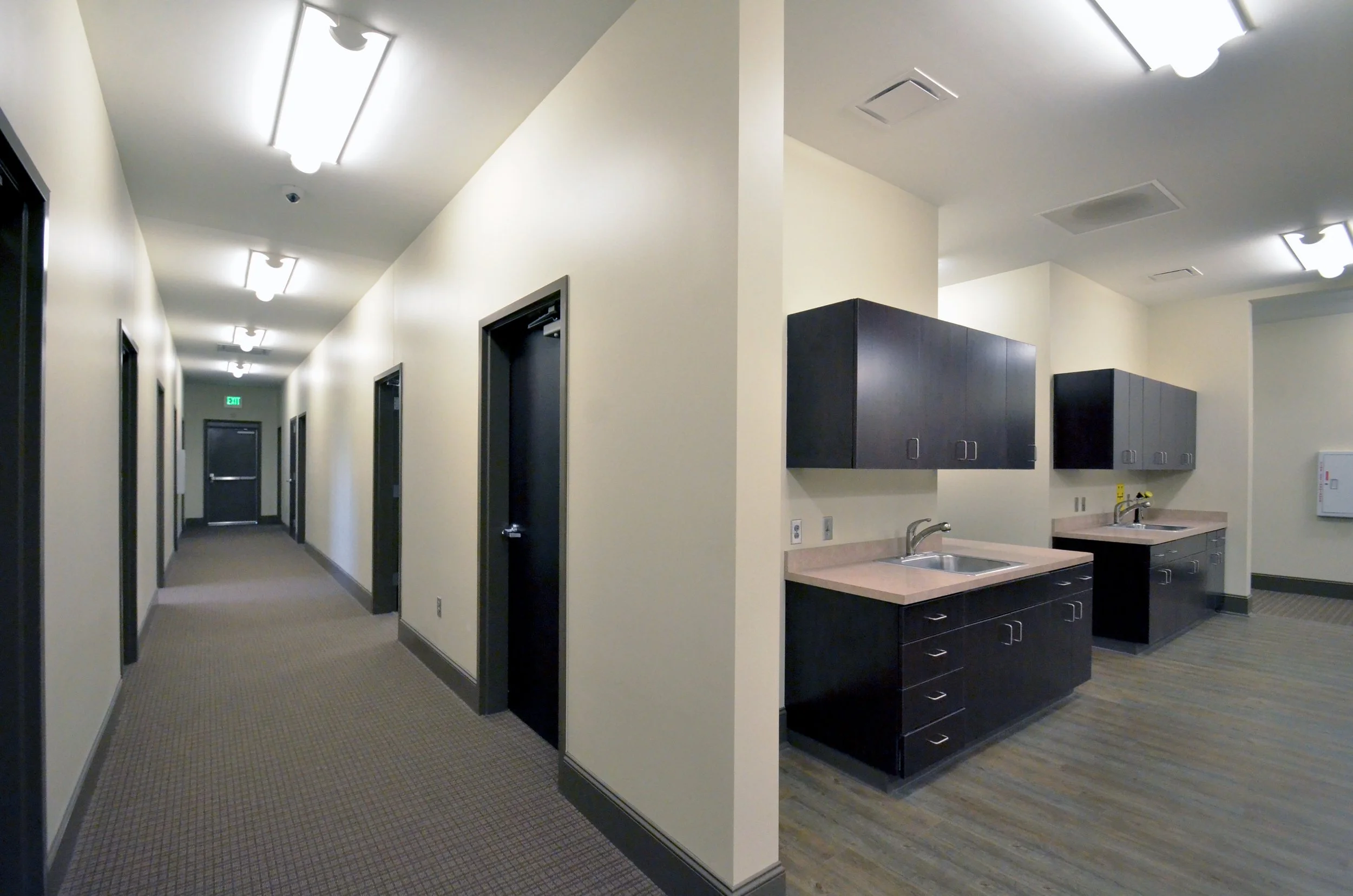 A hallway with multiple black doors on each side, beige walls, and ceiling lights. The flooring is carpeted. To the right, there is a kitchenette with black cabinets, sinks, and a countertop.