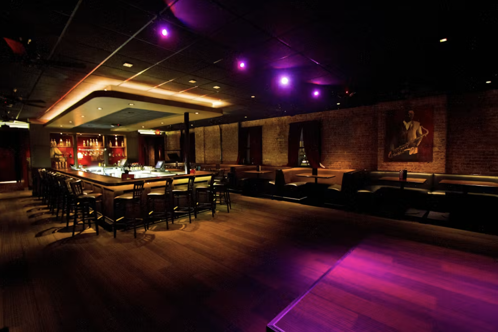 Empty dimly lit bar with a curved counter, high chairs, and wall art of a musician playing a saxophone, with exposed brick walls, purple ceiling lights, and wooden floors.