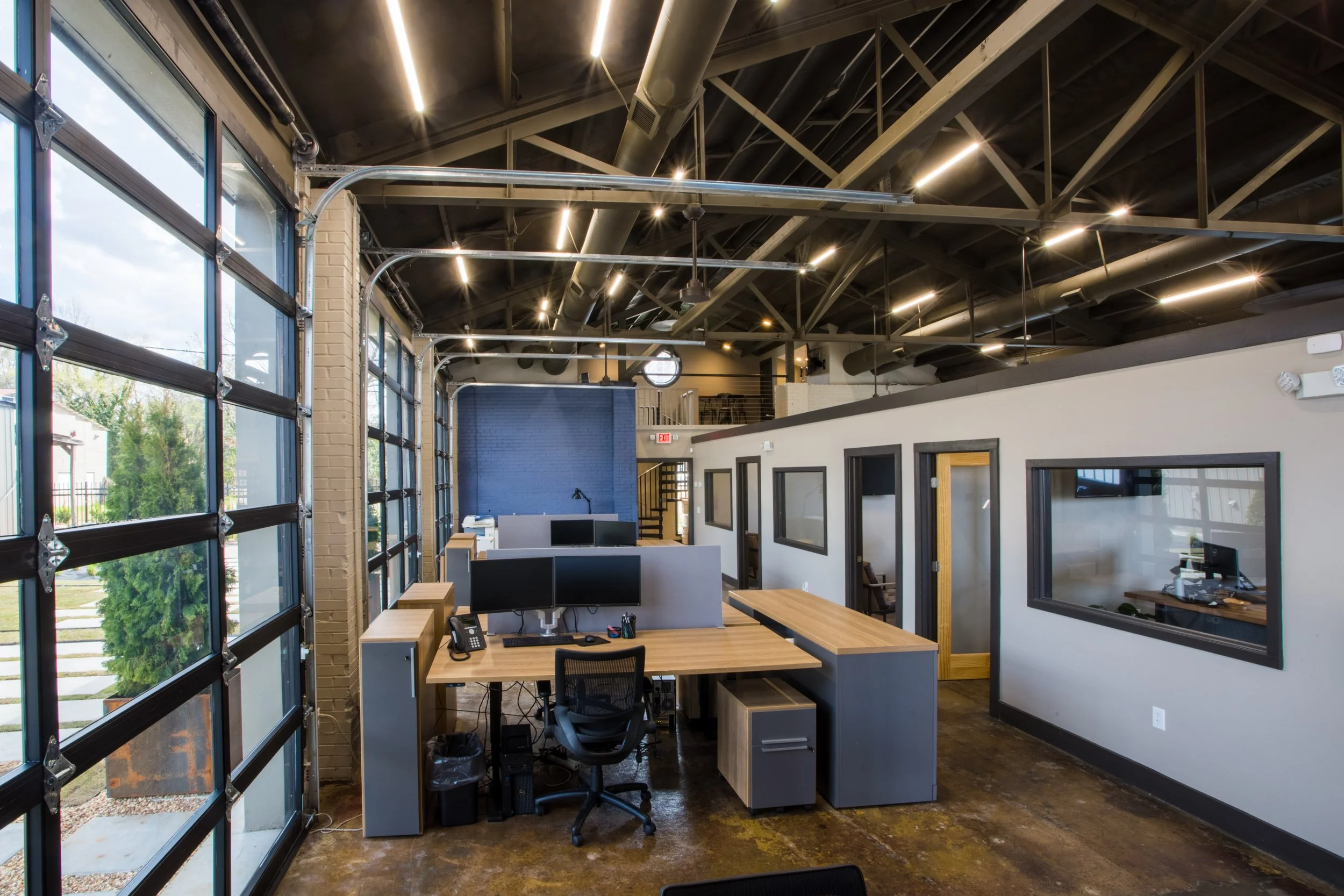 Modern office space with large windows, multiple computer workstations with monitors, chairs, and office supplies, exposed ceiling with ductwork and lighting, and a brick wall painted blue.