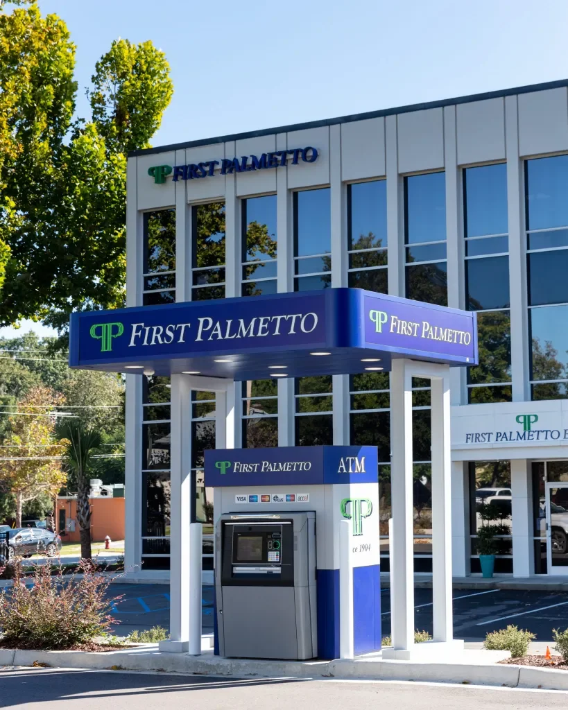 A small bank branch building for First Palmetto with an outdoor ATM, blue signage, and a modern glass exterior, surrounded by greenery and parking spaces.