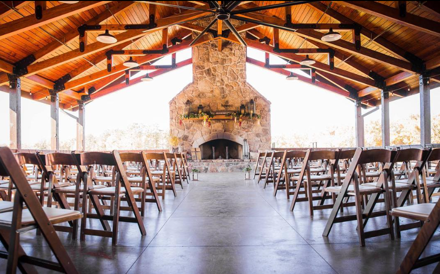 Indoor event space with wooden chairs arranged in rows facing a stone fireplace, under a high wooden ceiling with black structural beams and metal light fixtures.