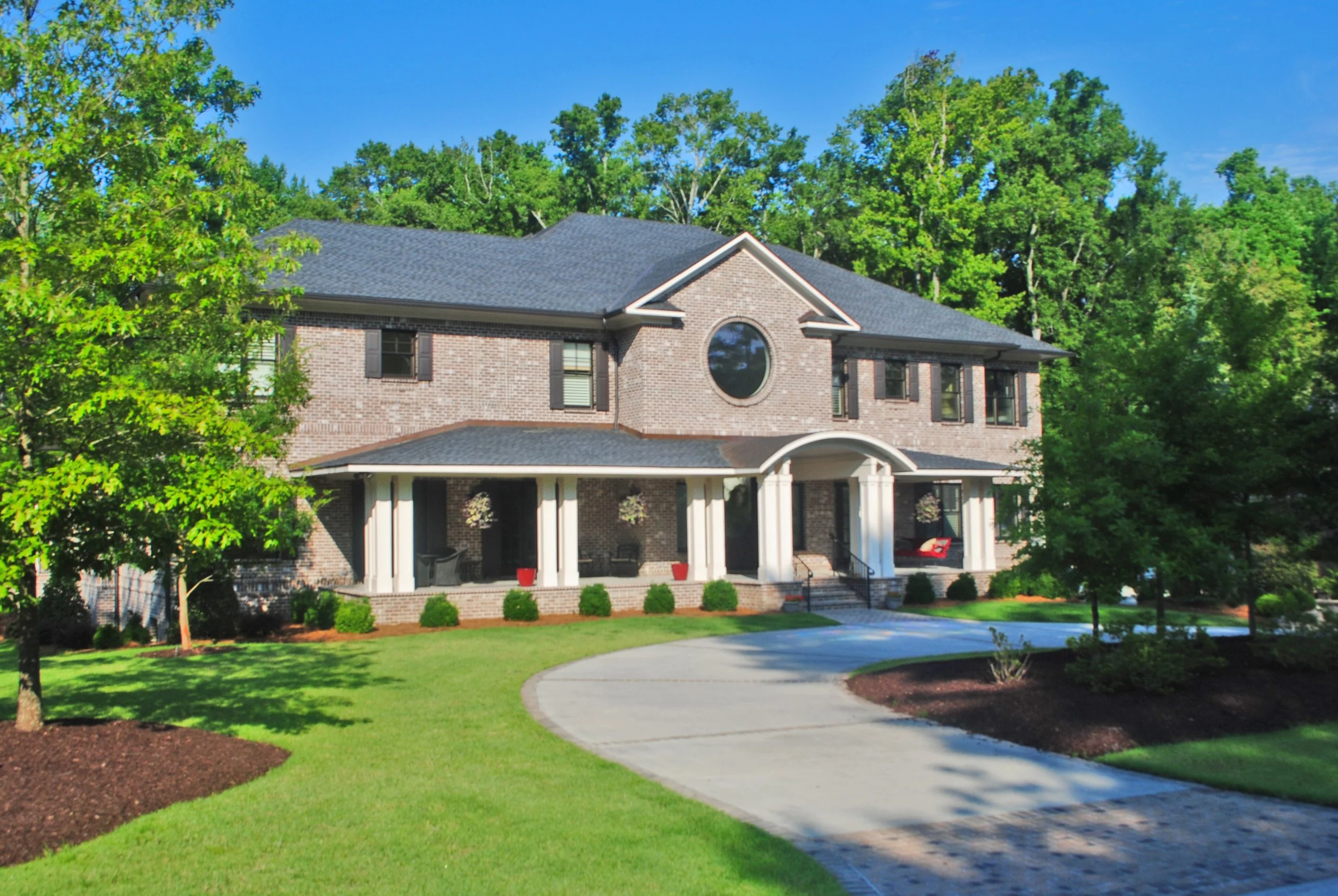 A two-story brick house with a dark shingle roof and a circular window in the front facade, surrounded by green trees and a landscaped yard with a curved concrete driveway.