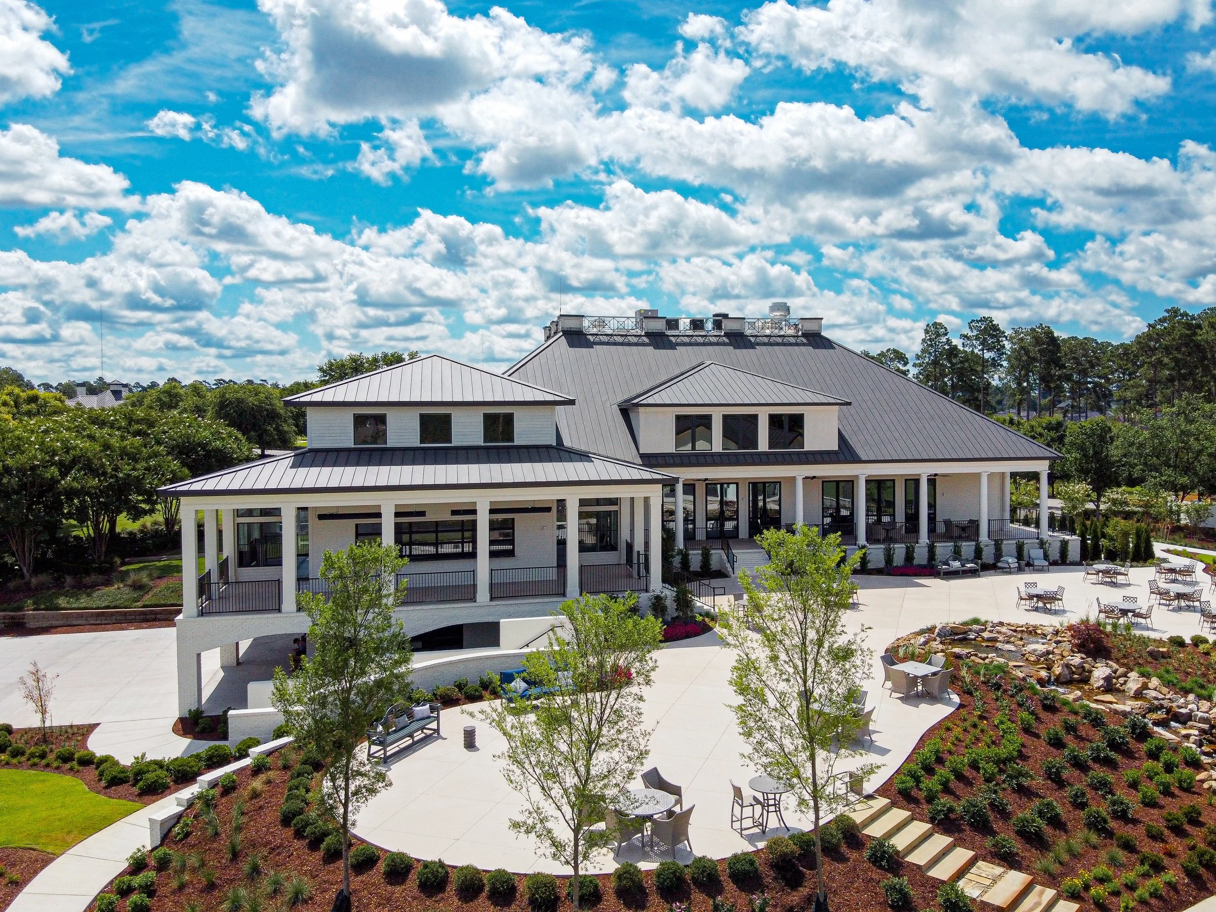 A large house with a metal roof, multiple levels, and wrap-around porch, surrounded by a landscaped yard with trees, bushes, and seating areas, under a partly cloudy sky.