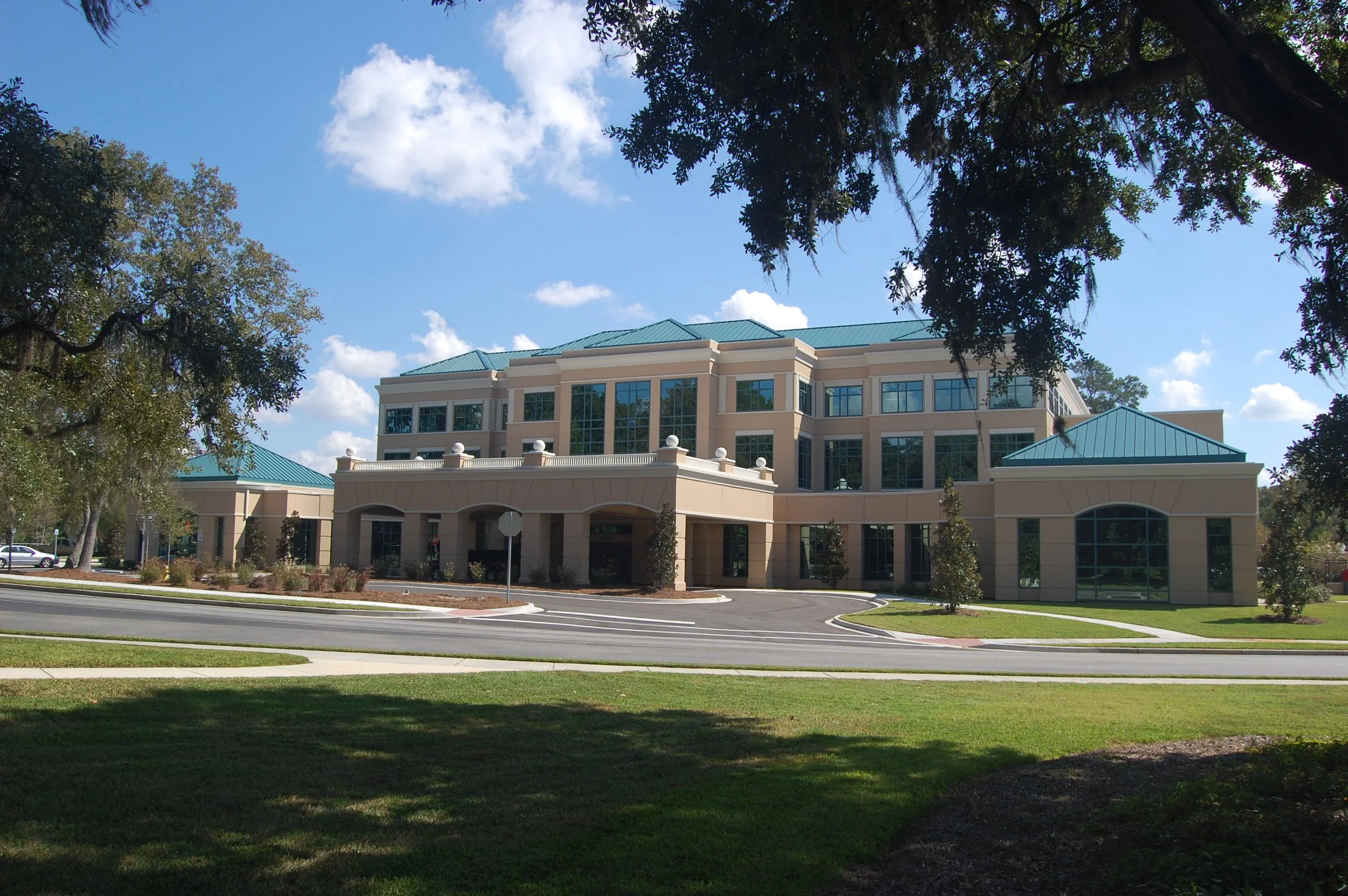 A large modern building with a pastel yellow and cream exterior, multiple large glass windows, and a teal metal roof. It is surrounded by a parking lot, landscaped greenery, and tall trees, visible under a blue sky with scattered clouds.