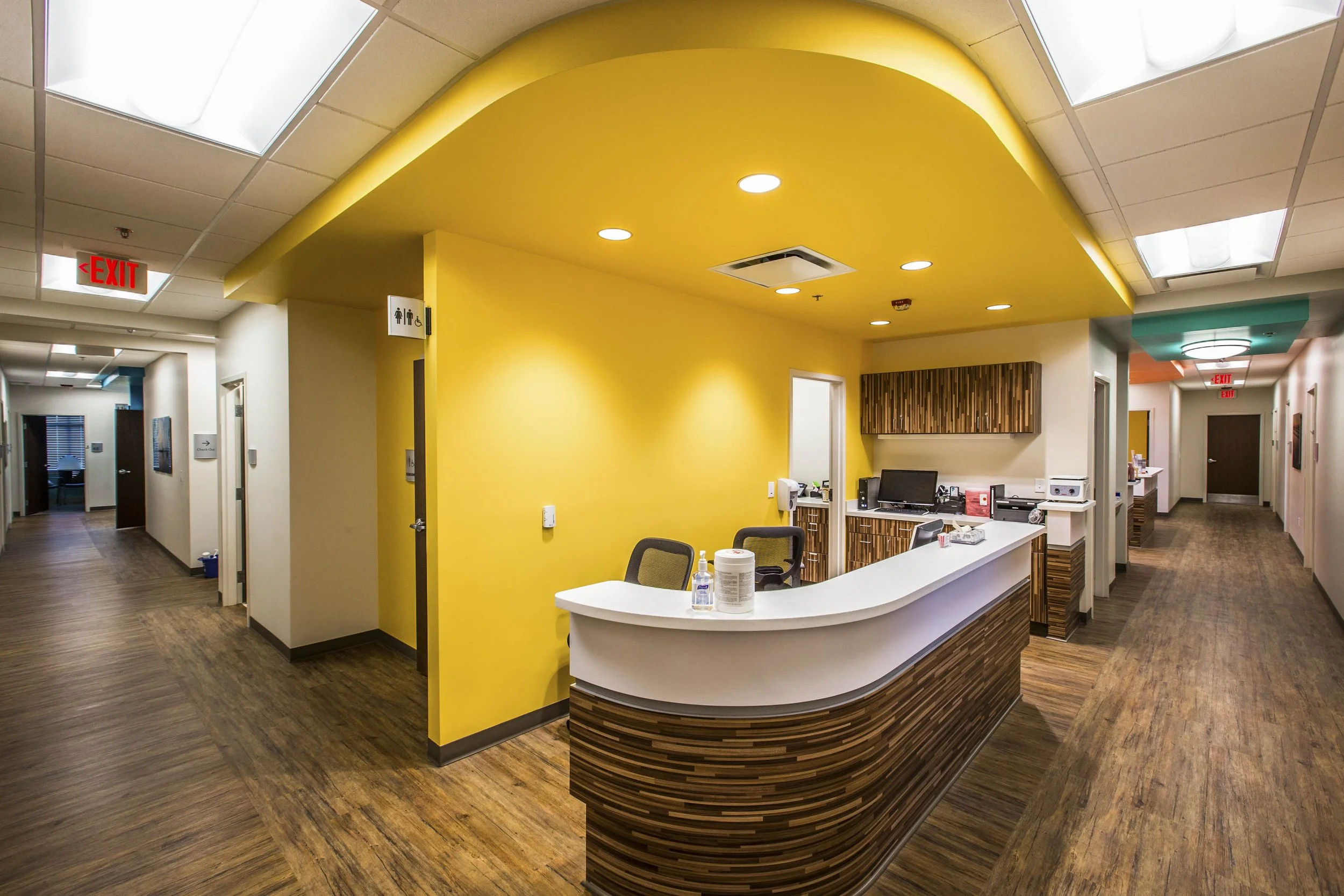 Interior of a medical office or clinic with a yellow accent wall behind a curved reception desk. The desk has a wood finish, and several office supplies are visible on it. The ceiling has large white panels with lighting, and signs for restrooms and 