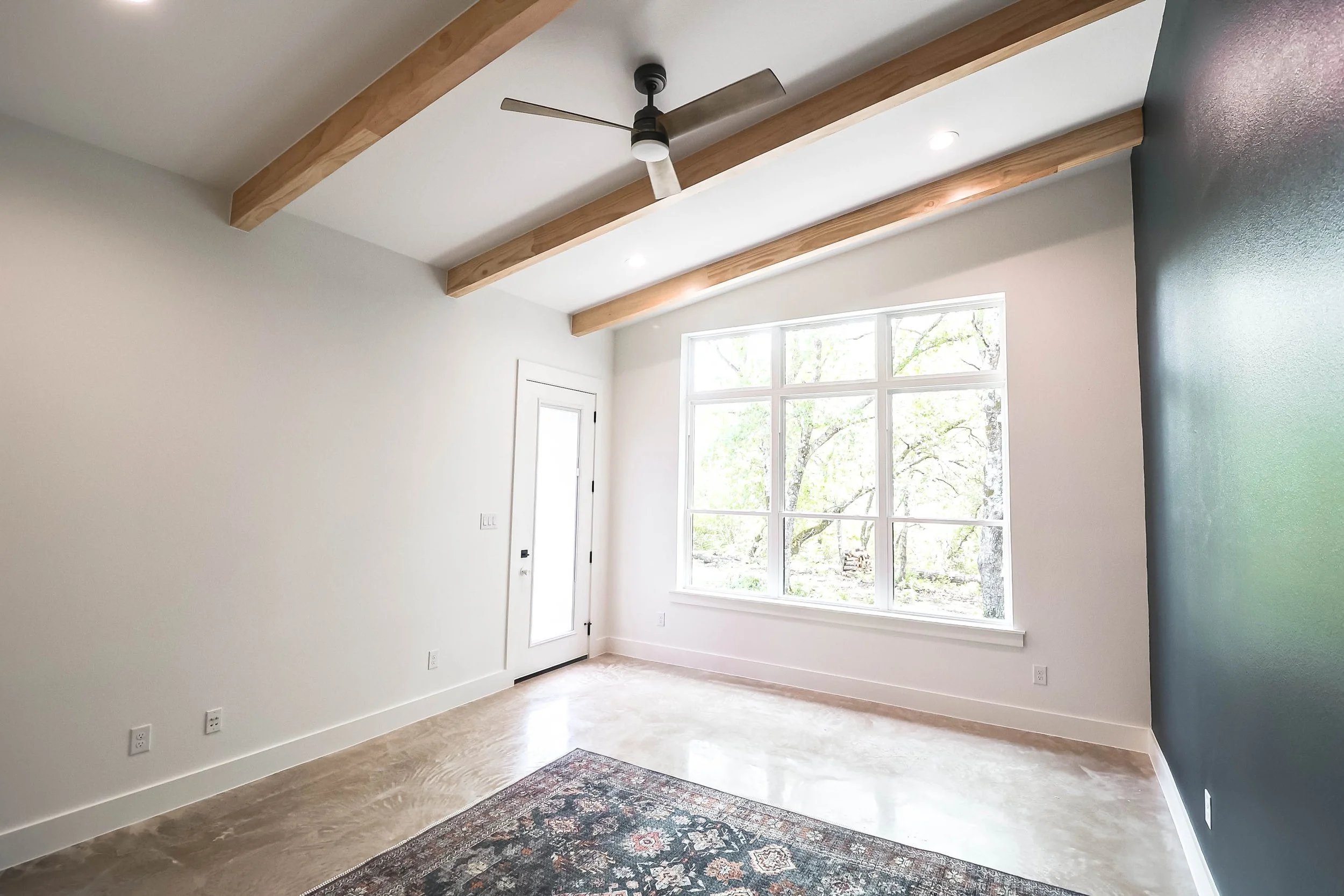 Empty room with large window, white walls, and wooden beams on ceiling.