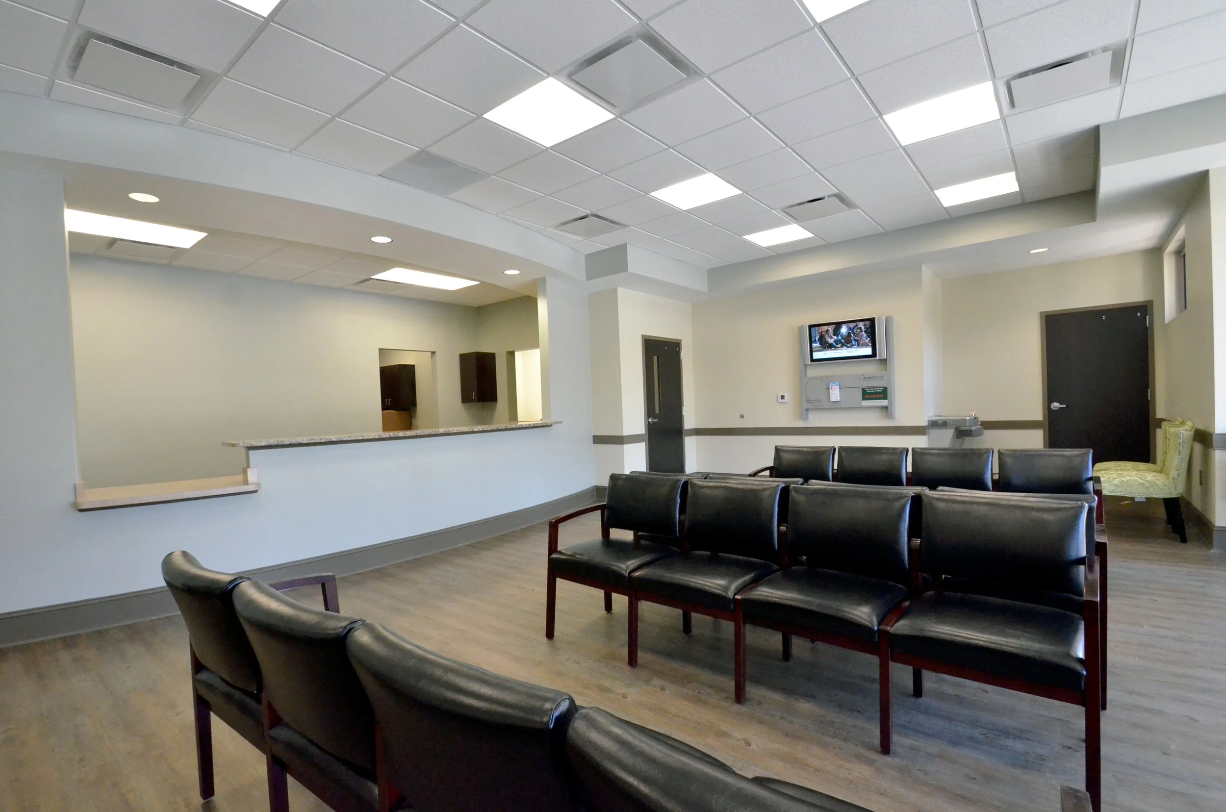 An empty medical waiting room with black chairs, a wall-mounted television displaying a news broadcast, and a reception counter with a granite top. The room has a dropped ceiling with fluorescent lighting and cream-colored walls.